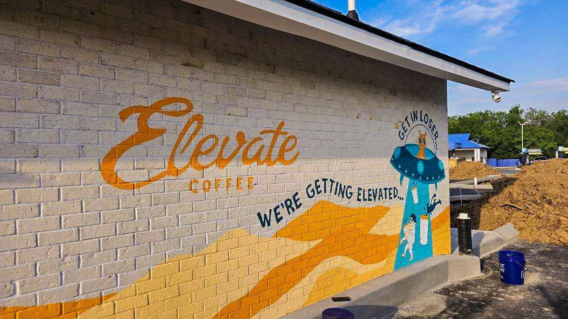 Colorful mural on a brick wall with the words 'Elevate Coffee' and a cartoon astronaut with a dog inside a spaceship, with the phrase 'Get in Loser, We're Getting Elevated'.