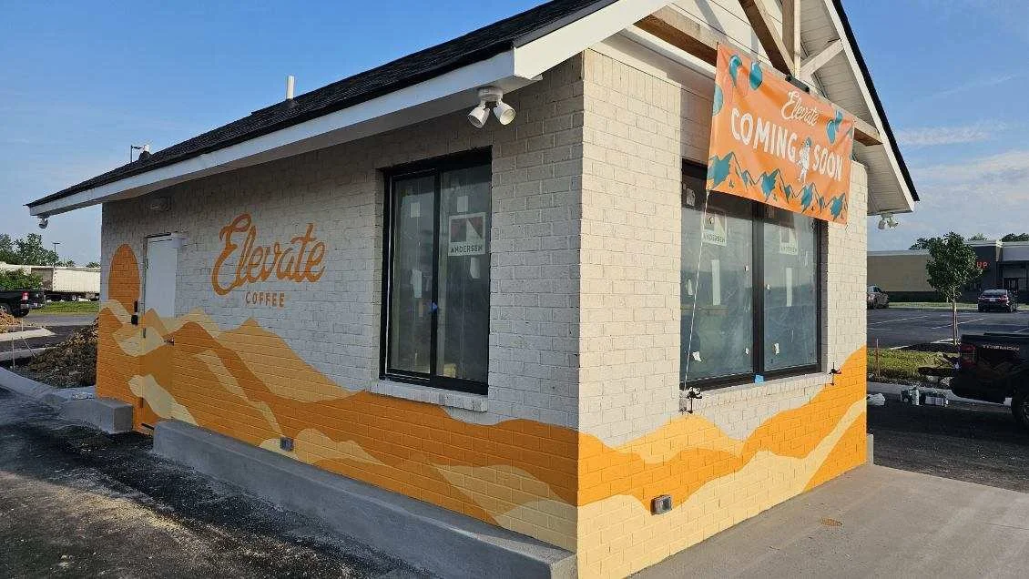 A brick building with a mural of mountains painted in shades of orange and yellow. The building has a sign that reads 'Elevate Coffee' and an orange banner with 'Coming Soon.' The windows are large, and the building is located in a parking lot.