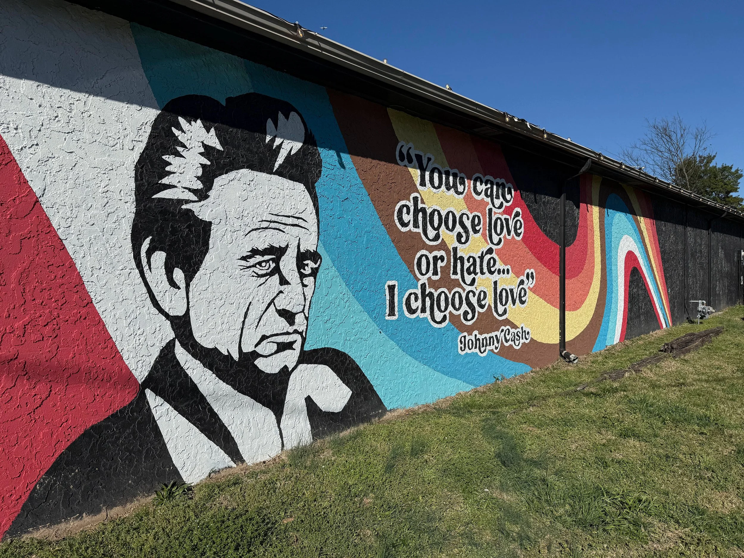 Colorful mural on a building wall featuring a black and white portrait of a woman with short hair and a serious expression. Next to her is a quote by Johnny Cash, reading 'You can choose love or hate... I choose love,'' with a rainbow-colored design 
