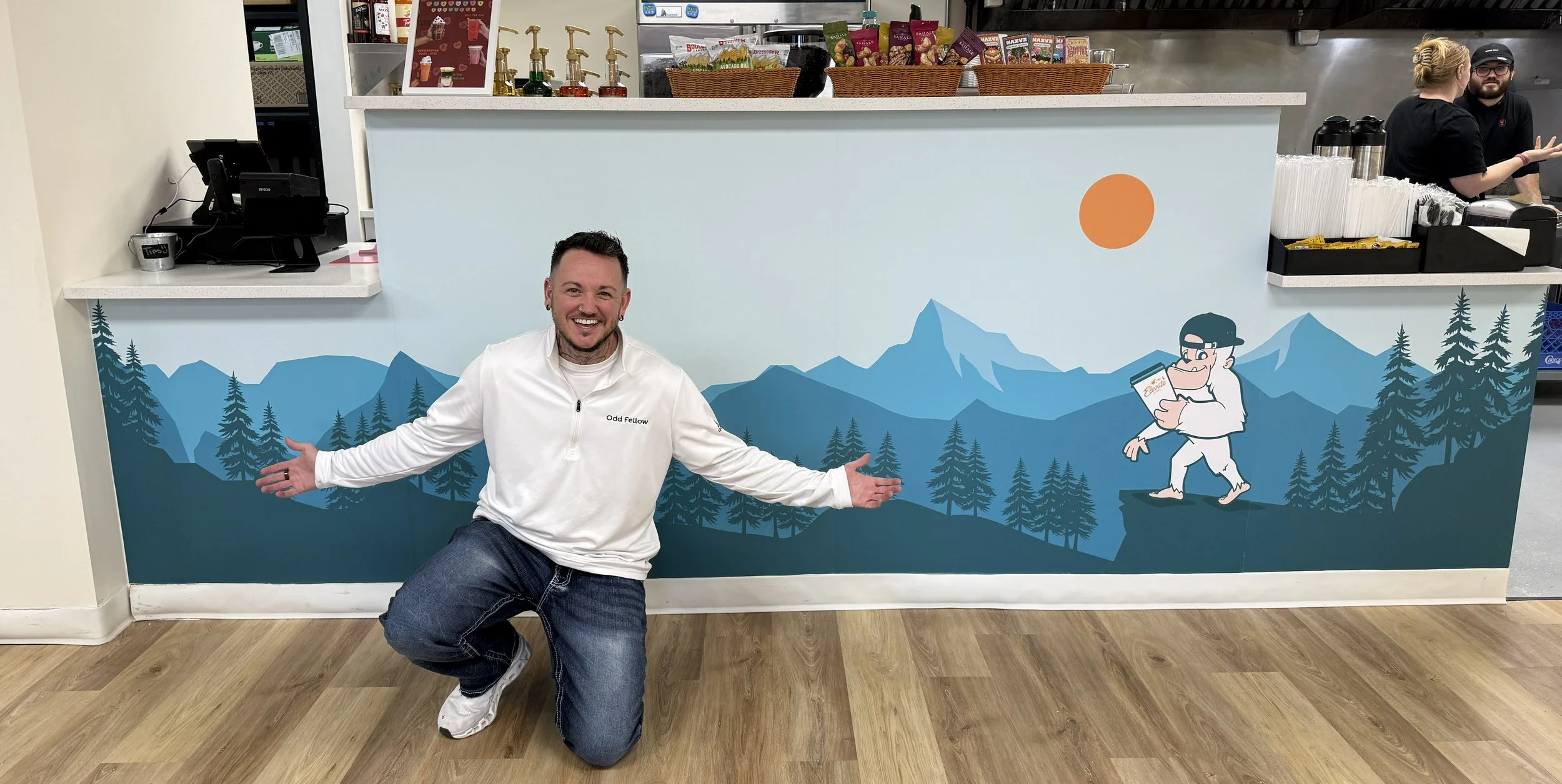 Man kneeling in front of a mountain and forest mural inside a cafe, smiling and gesturing with arms outstretched.