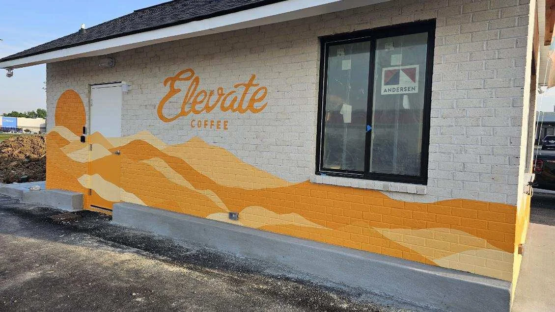 Exterior of a coffee shop with a mural of stylized orange and yellow mountain ranges and the words 'Elevate Coffee' on a white brick wall.