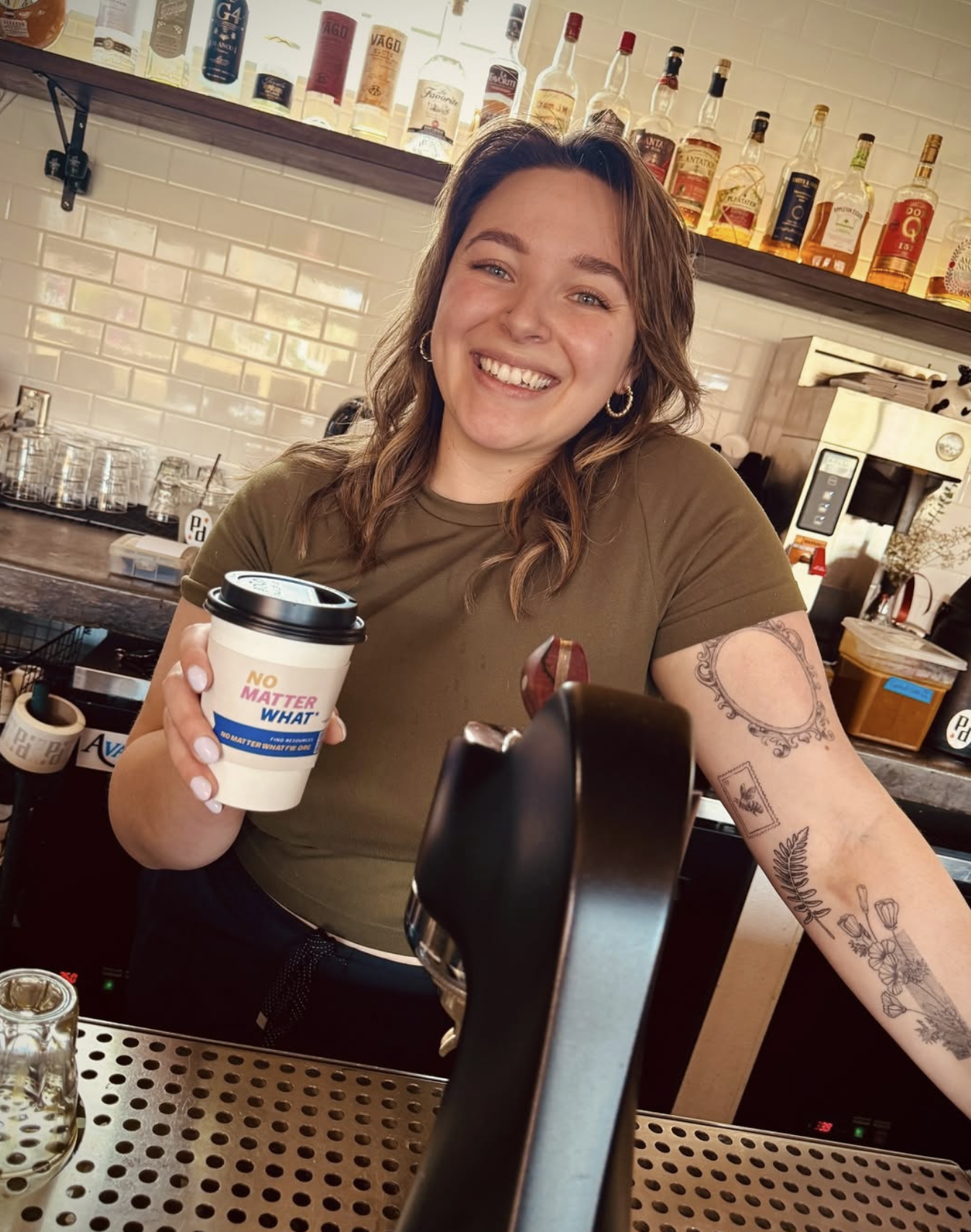 A smiling woman behind a bar counter holding a coffee cup that says "No Matter What", with a tiled wall and bottles of cafe supplies in the background.