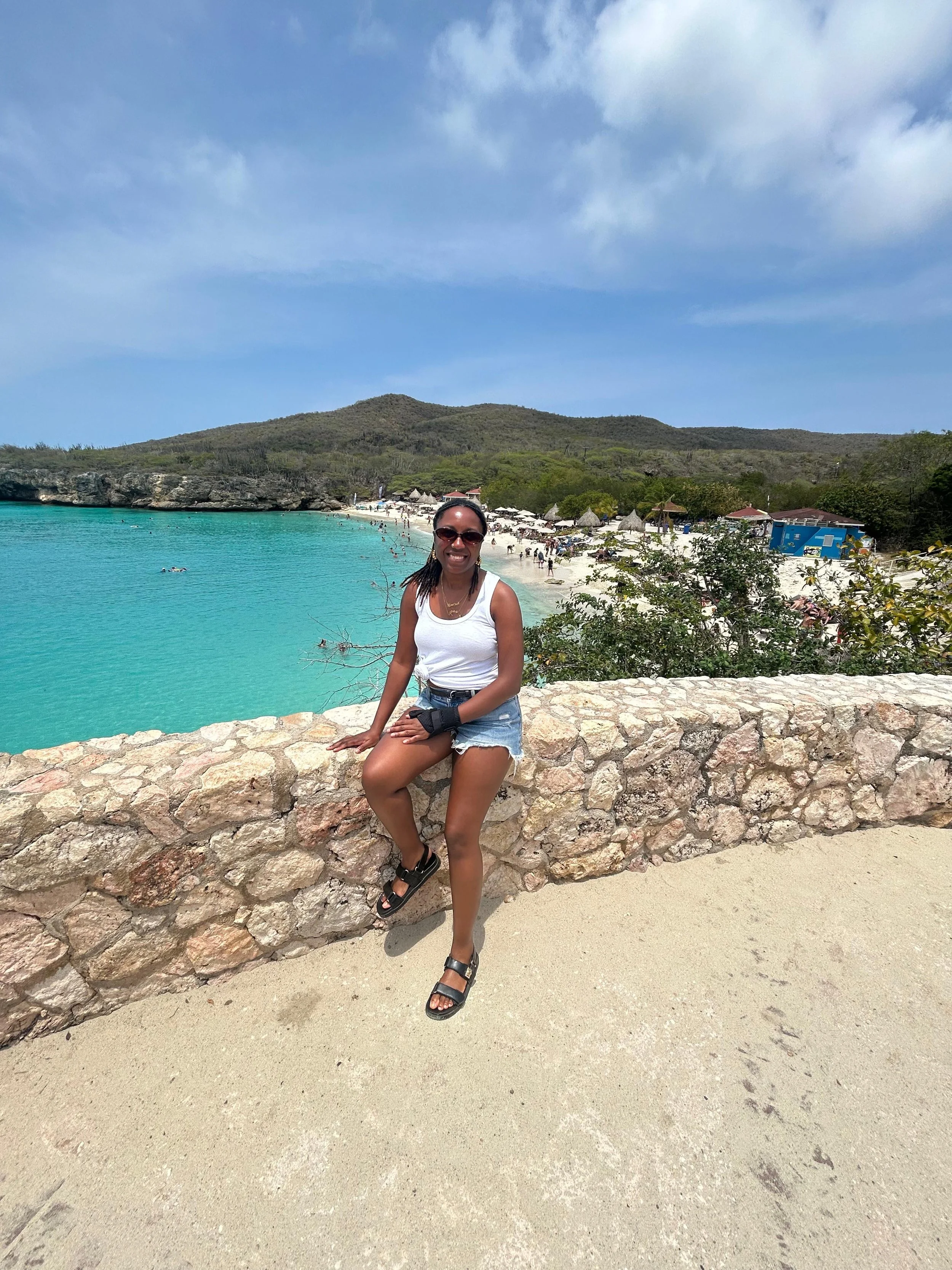 Jessica Thomas sitting on a low stone wall at the edge of a beach in Curaçao, turquoise water behind her.