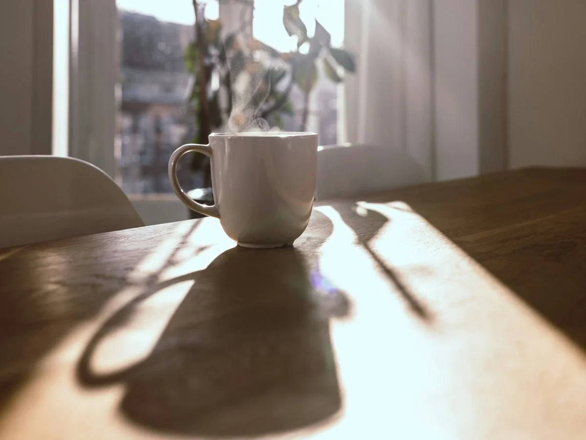 A steaming white coffee mug on a wooden table, with sunlight casting shadows and a window with curtains in the background.