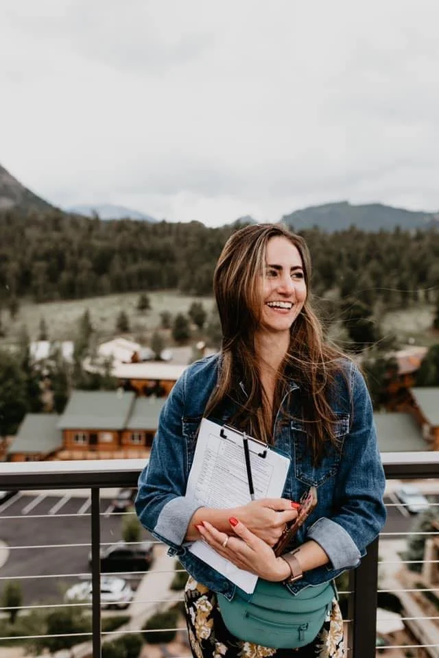 Nicole Fauls, Owner of Urban Allure Events, standing on a balcony holding a clipboard and smartphone, with a Colorado landscape of mountains and trees in the background.