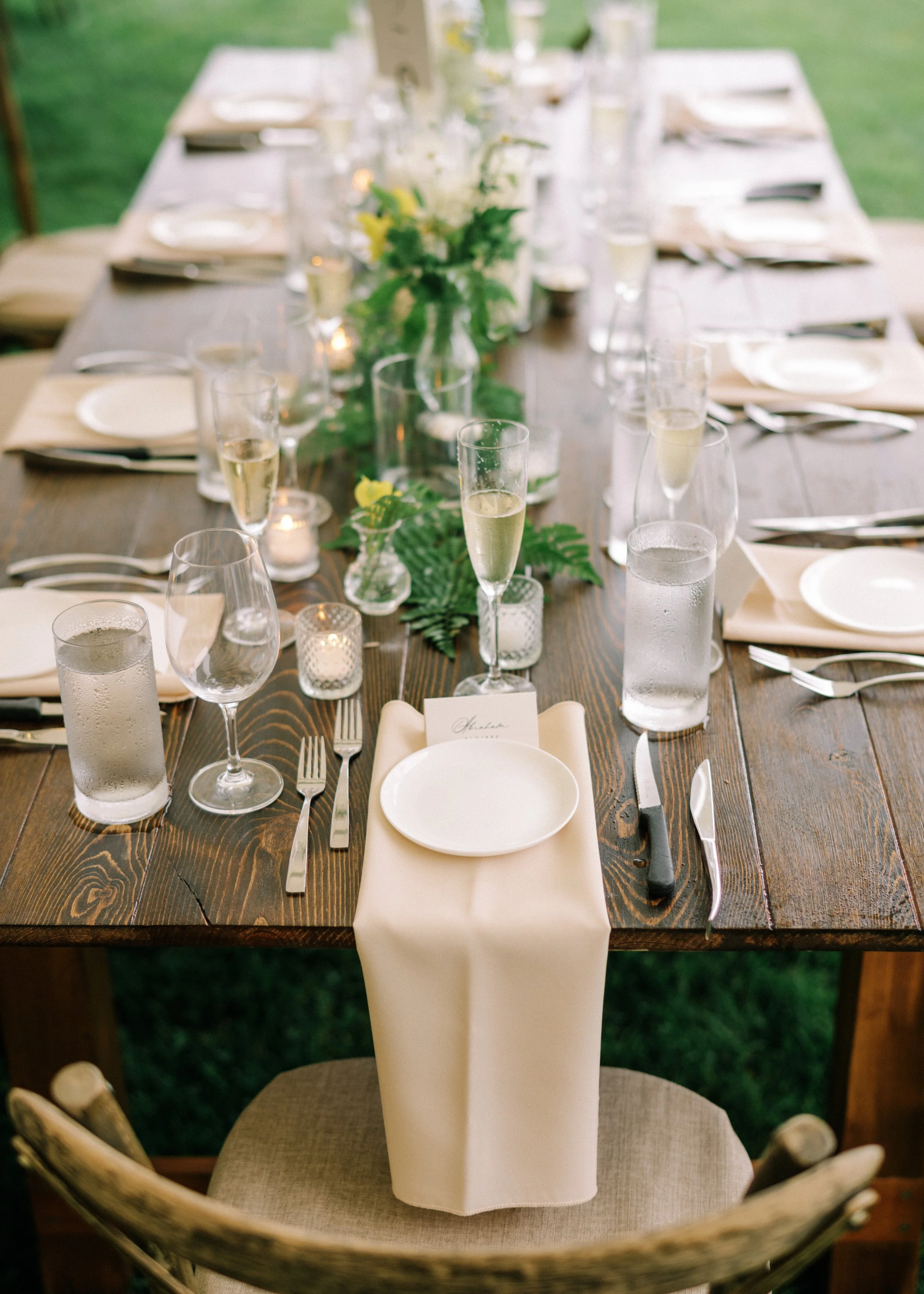 A formally set outdoor dining table with a dark wooden surface, white plates, silverware, champagne glasses, water glasses, small candles, and green floral arrangements down the center, with chairs surrounding it, on a grassy area.