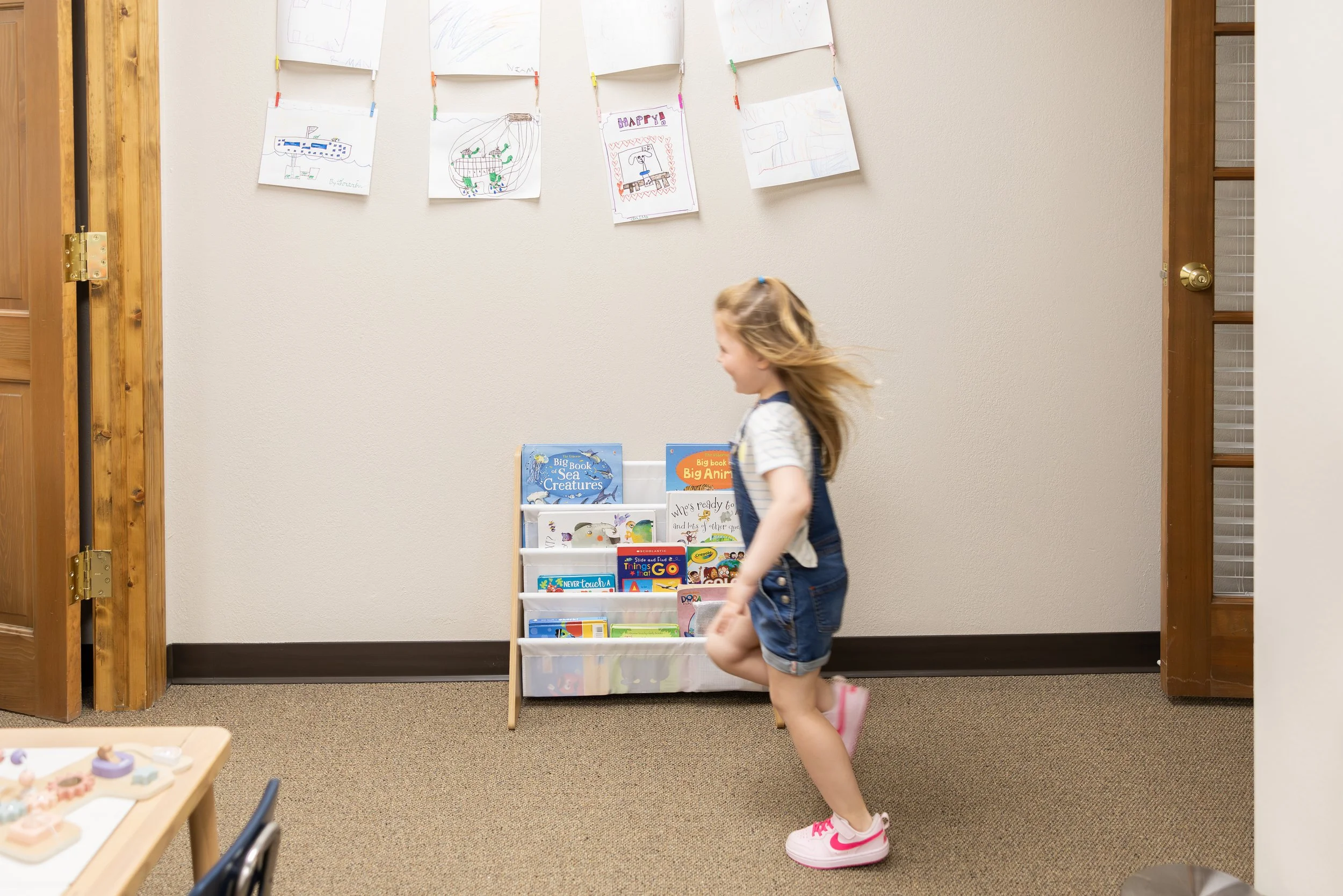 A young girl with blonde hair in a ponytail, wearing a striped shirt, denim shorts, and pink and white sneakers, is running past a bookshelf filled with children's books inside a room with beige walls and brown carpeting. Artworks are hanging on the 
