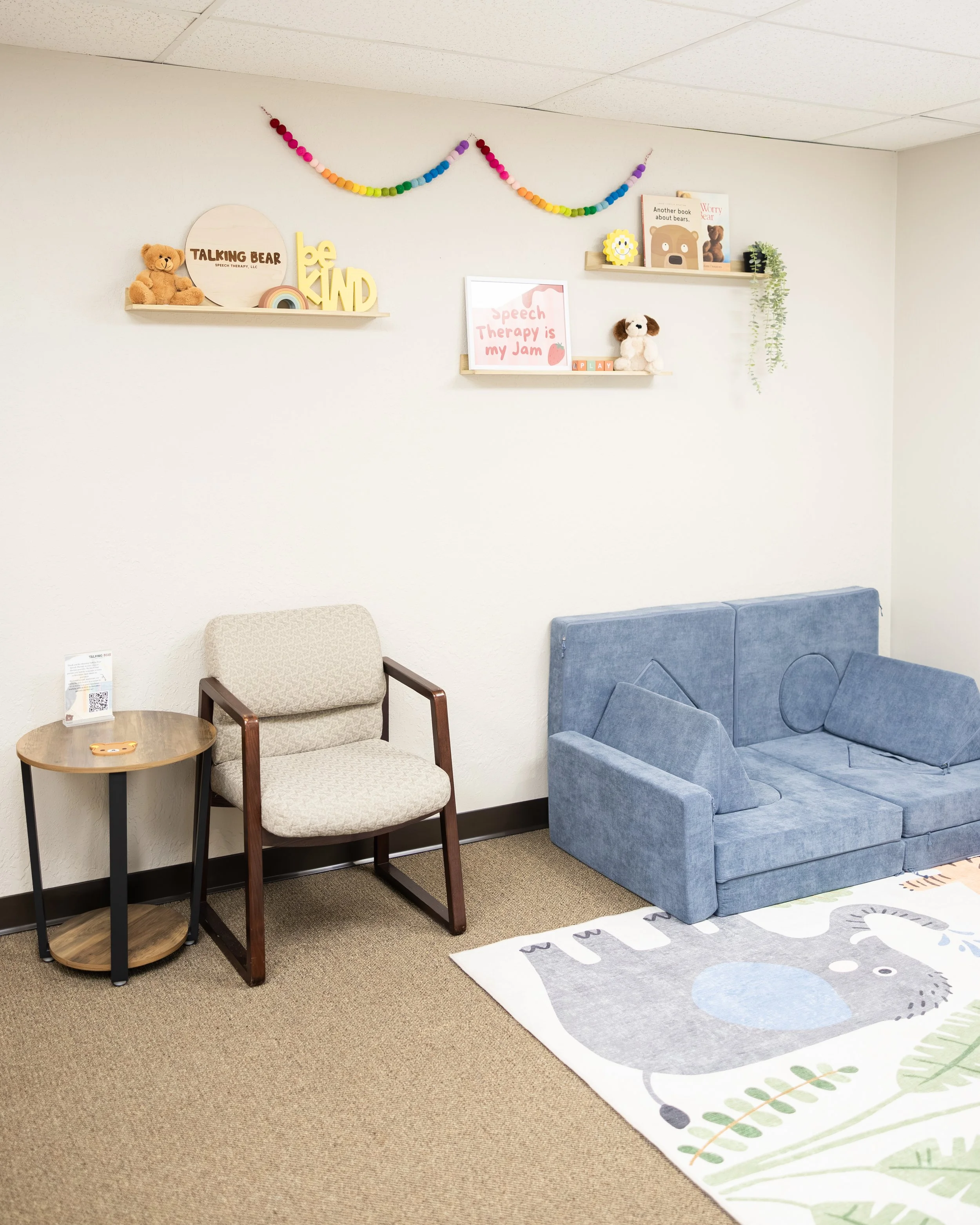 A cozy waiting room with beige and blue seating, decorated with a children's themed rug and wall shelves holding stuffed bears, children's books, and a rainbow garland.