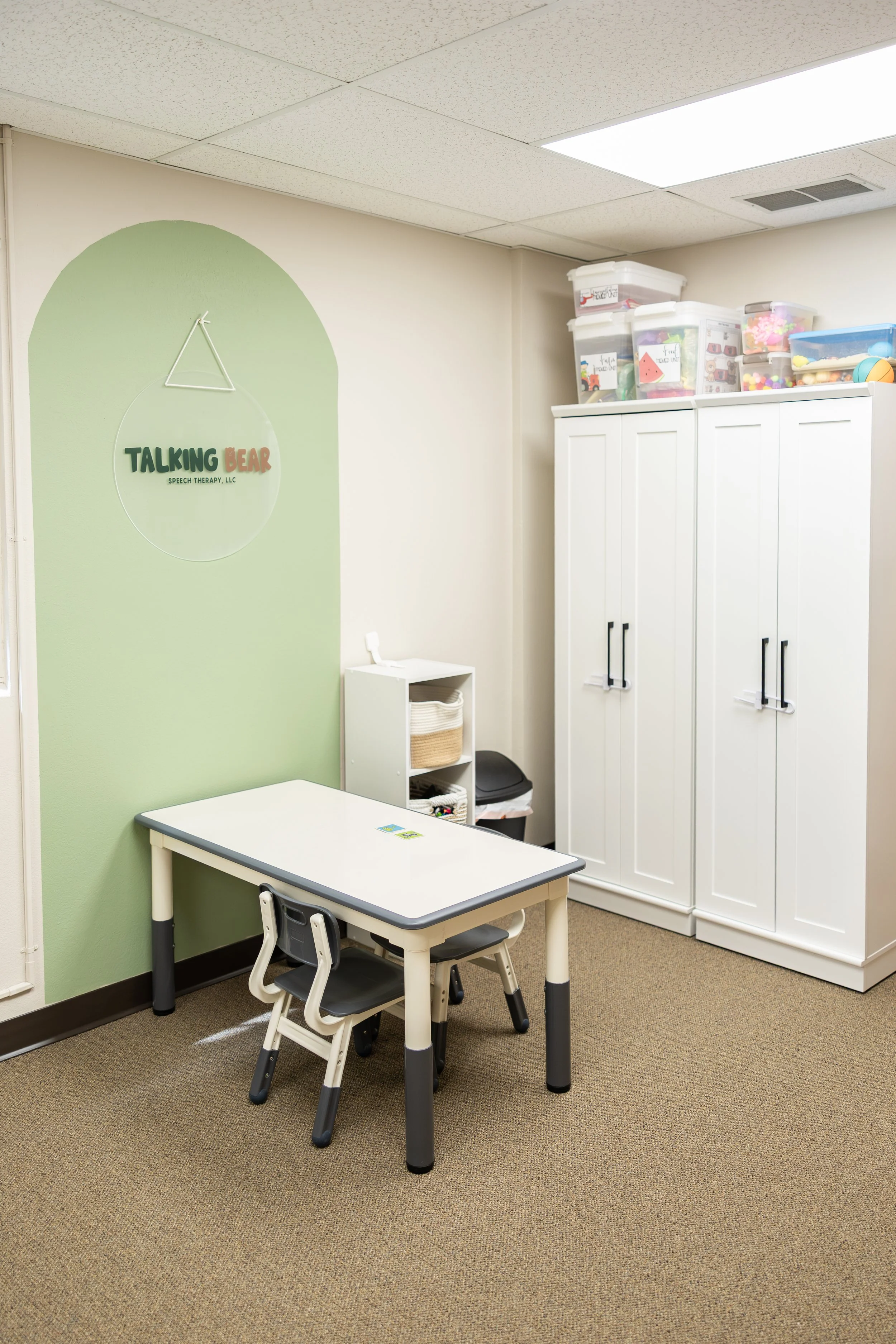 A small room with a green wall, a white table with two small black chairs, a white storage cabinet, and plastic containers filled with toys, with a sign that says "Talking Bear Speech Therapy" on the wall.