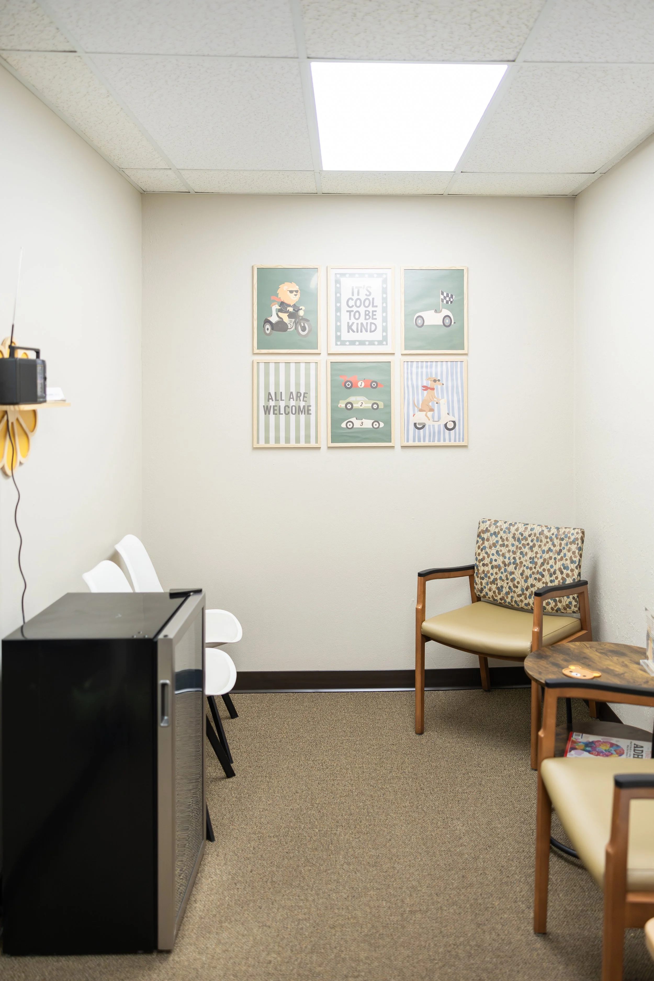 A small waiting room with beige carpet, six chairs, a desk, a small cabinet, and framed pictures on the wall, including a sign that reads 'It's cool to be kind'.
