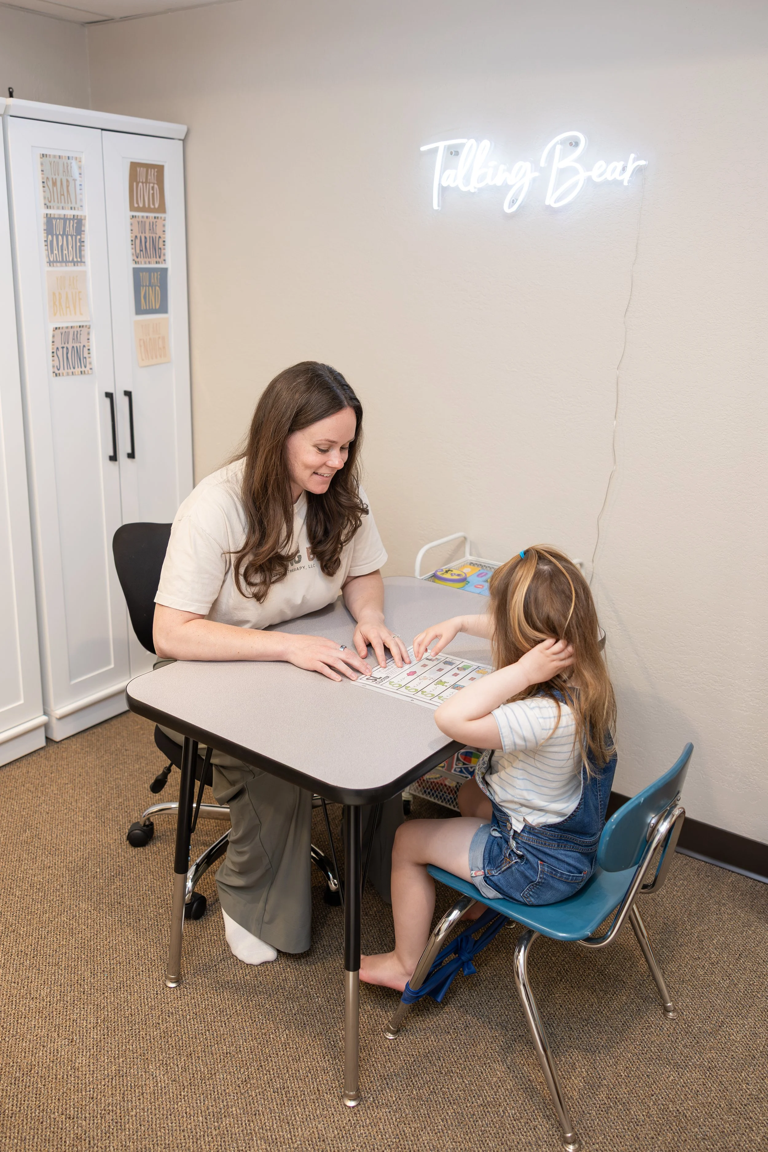 A woman sitting at a desk with a young girl, engaging in a learning activity. The woman is smiling and pointing at a worksheet, while the girl is sitting in a blue chair, touching her head. In the background, there is a white cabinet with motivationa