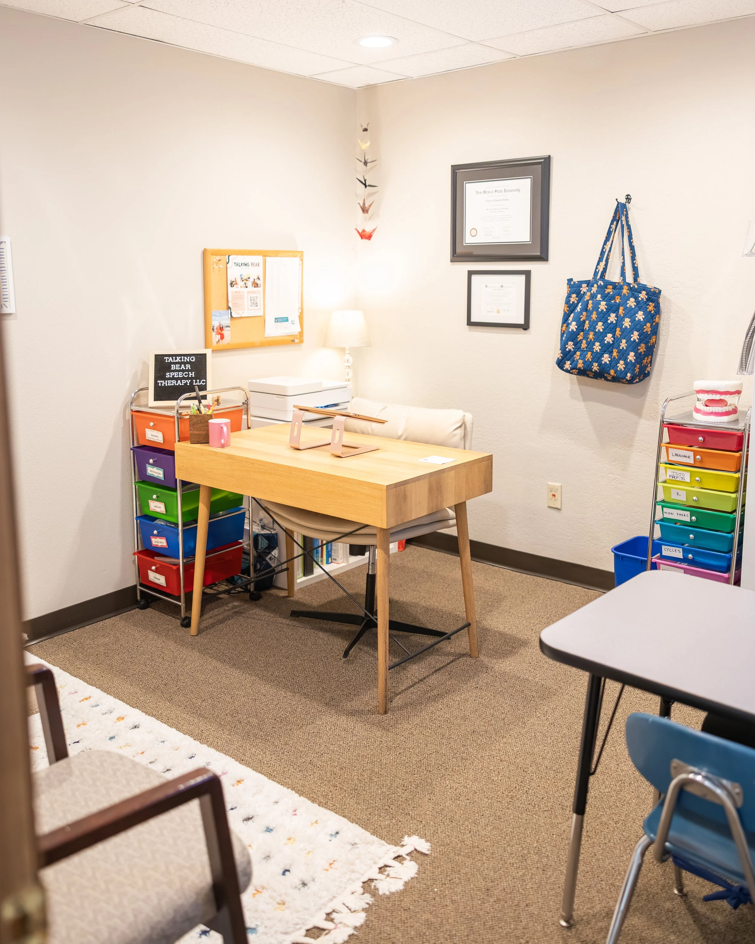 Office or therapy room with colorful storage drawers, a table with chairs, a wall with framed certificates, a hanging bag, and other decor.