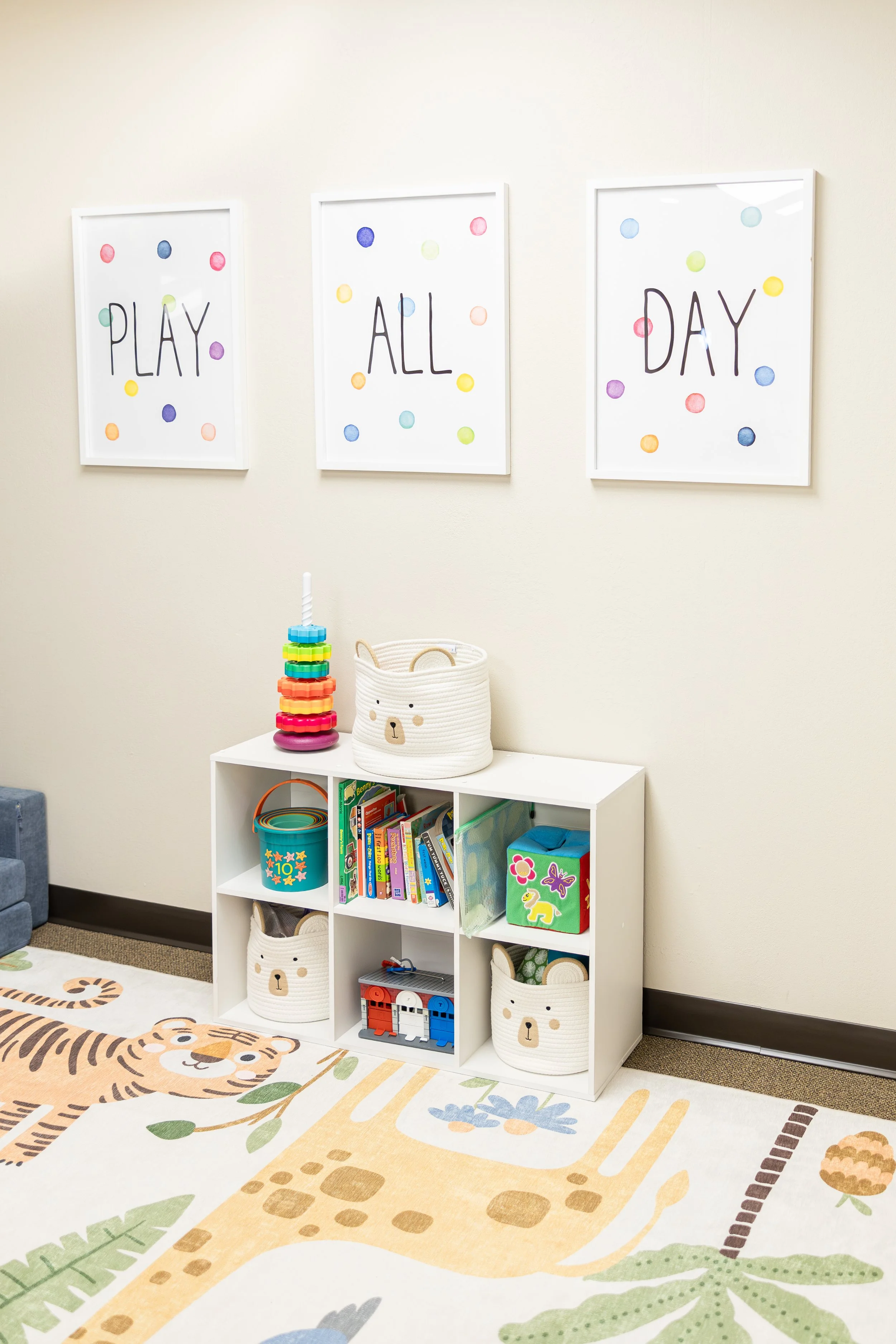 Decorative wall art reading 'Play All Day' with colorful polka dots, filled with children's toys, books, and a fun animal-themed rug in a children's playroom.