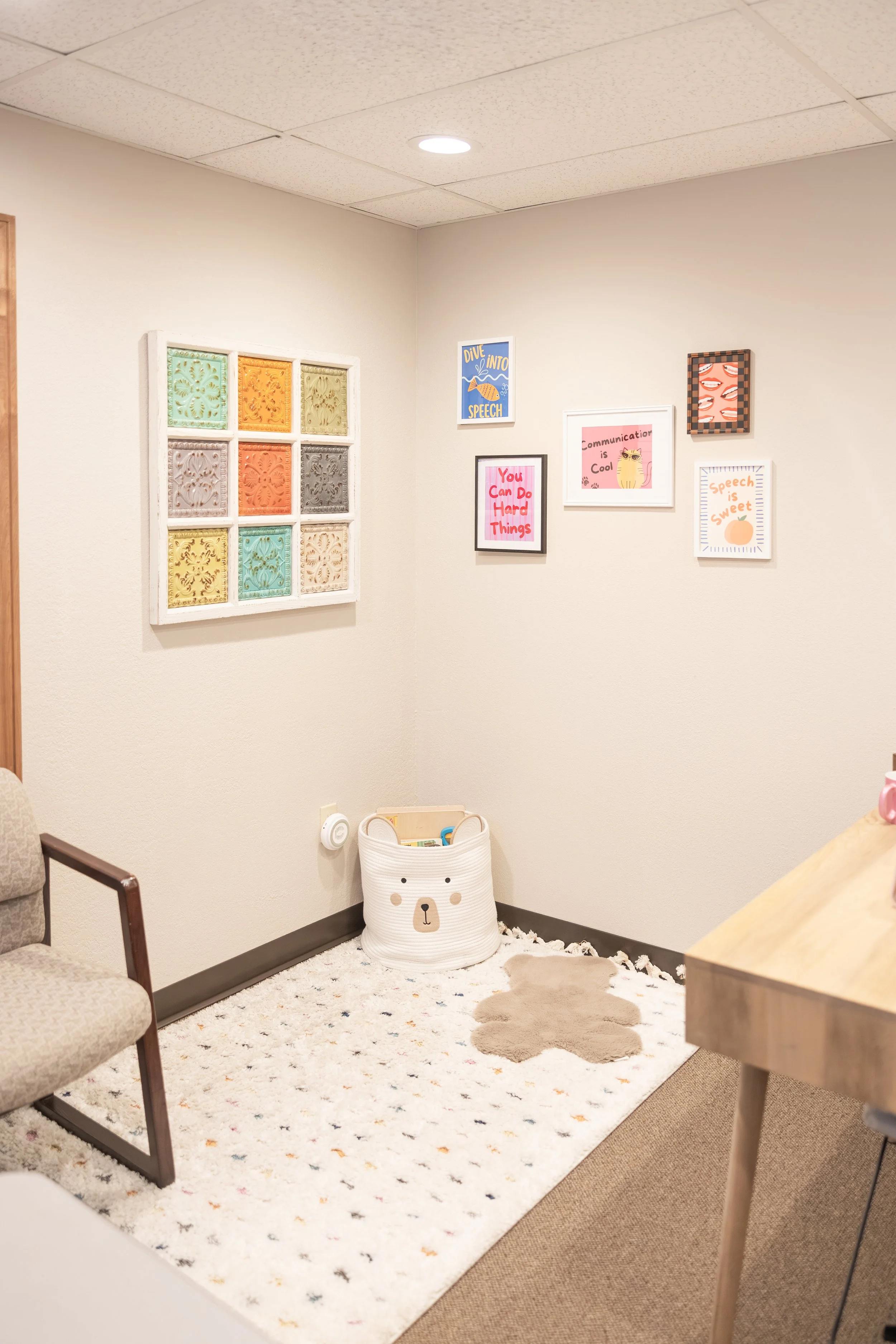 A cozy corner in a room with a framed collection of colorful decorative tiles on a cream wall, surrounded by various framed motivational and fun artwork about speech and communication. The corner has a plush white rug with multicolored dots, a white 