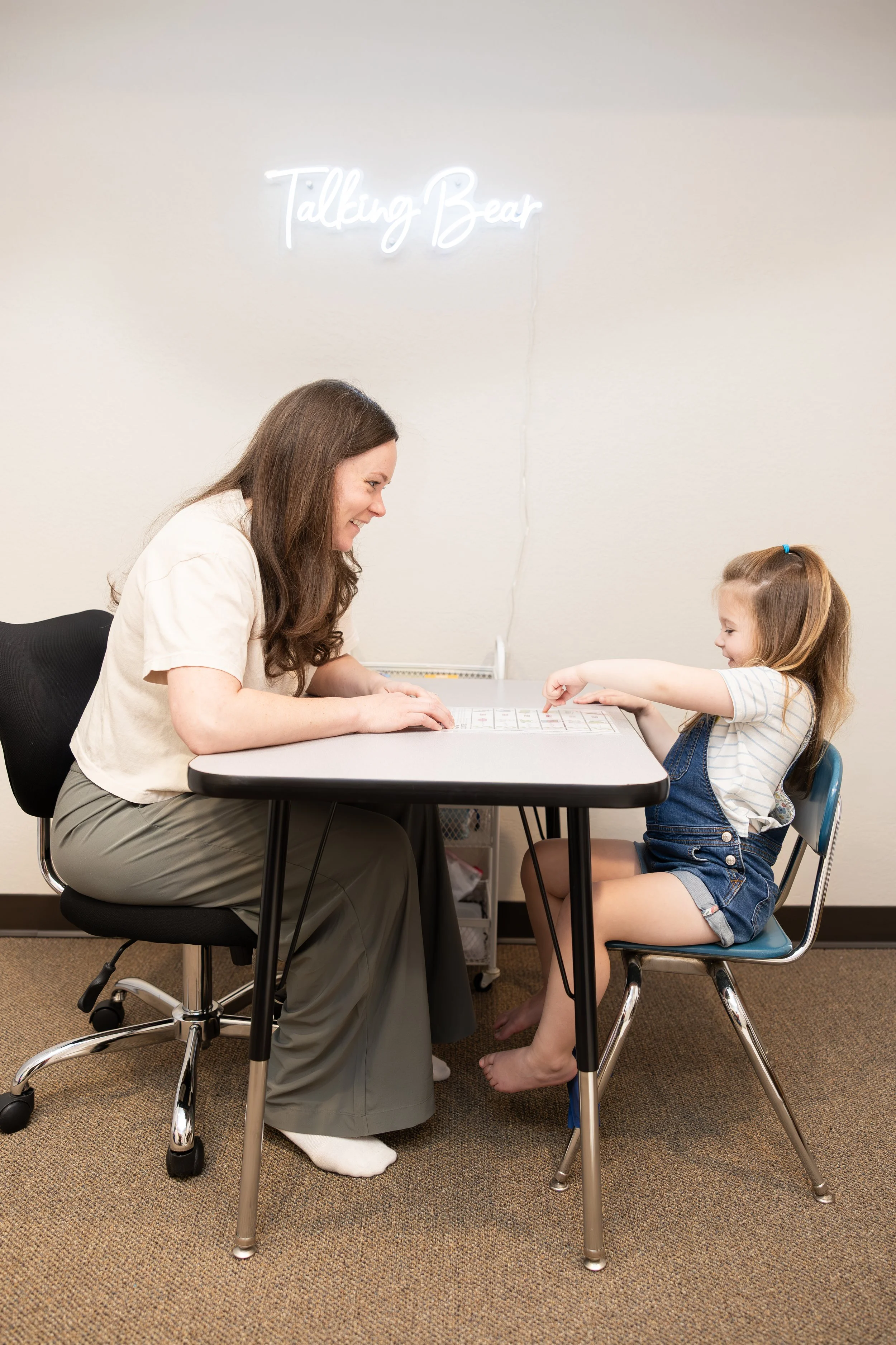 A woman and a young girl are sitting across from each other at a table, smiling and engaging in an activity together. The woman has long brown hair, is wearing a beige shirt and gray pants, and is sitting on a rolling office chair. The young girl has