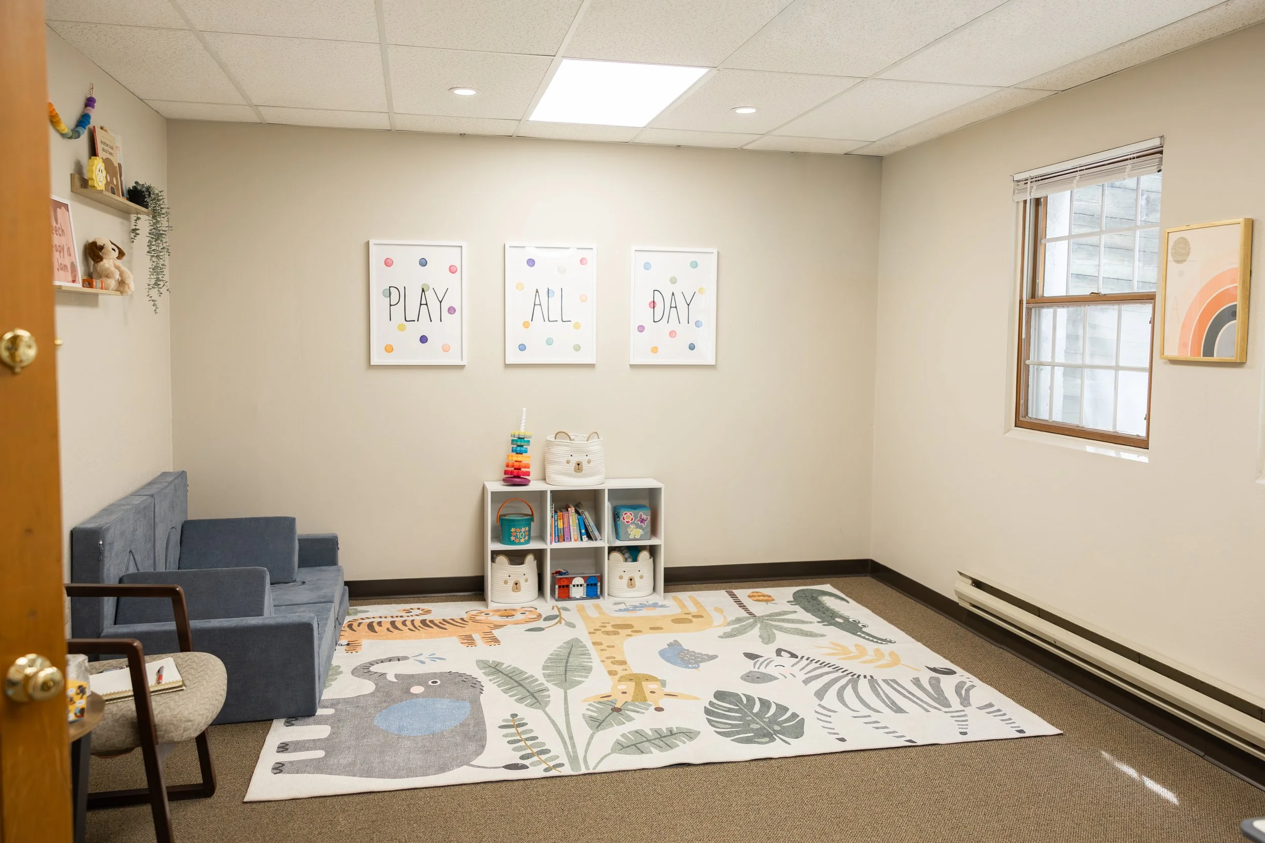 A children's playroom with beige walls, decorated with framed posters that say 'PLAY ALL DAY' and colorful dots. There are toy storage baskets shaped like bears, a small bookshelf with books and toys, colorful wall decorations, and a grey sofa. The f
