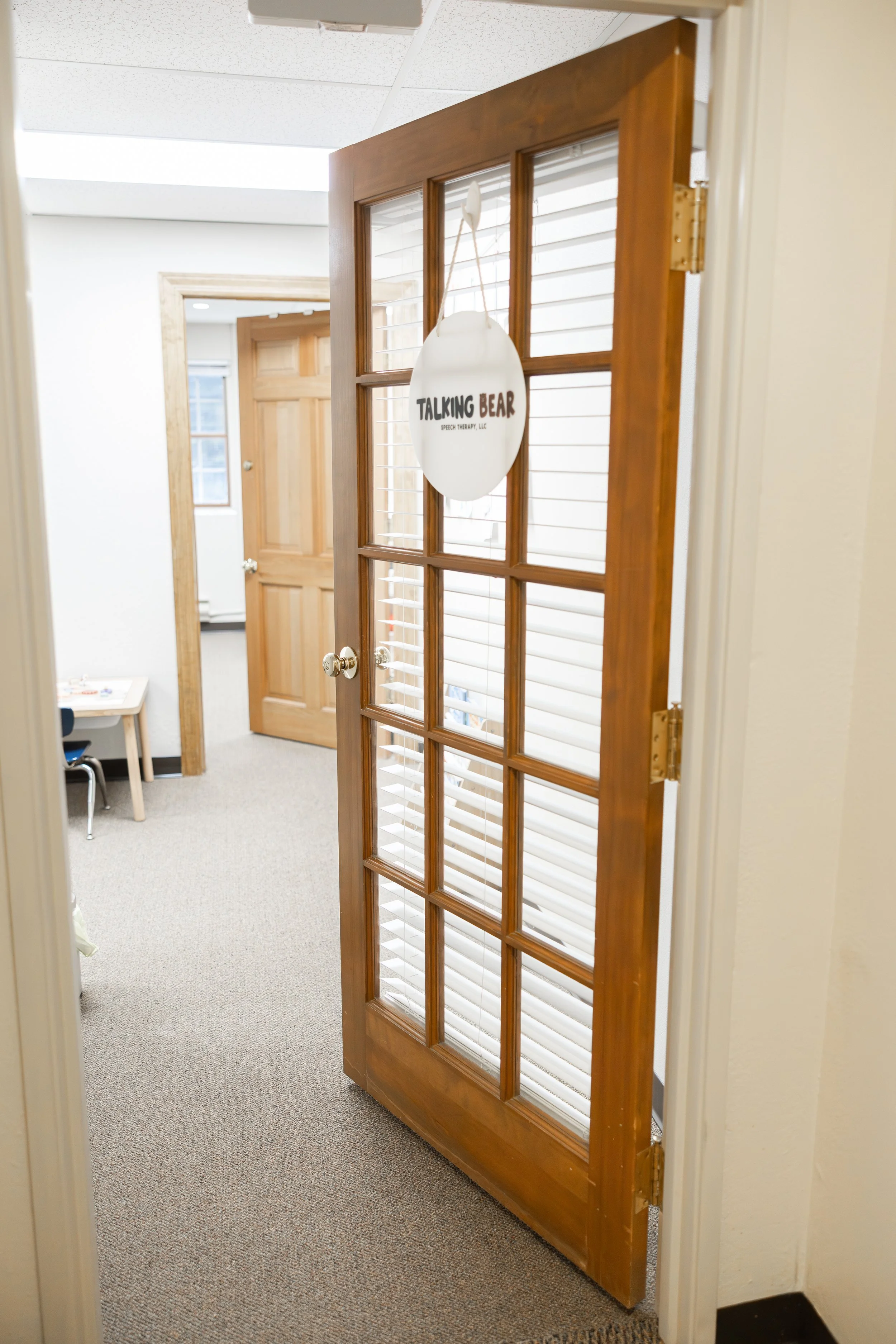 Open wooden door with glass panes leading into a small office or classroom, with a sign hanging on the door that reads "Talking Bear Speech Therapy LLC." Inside the room, there is a small table and a chair, with white window blinds behind.