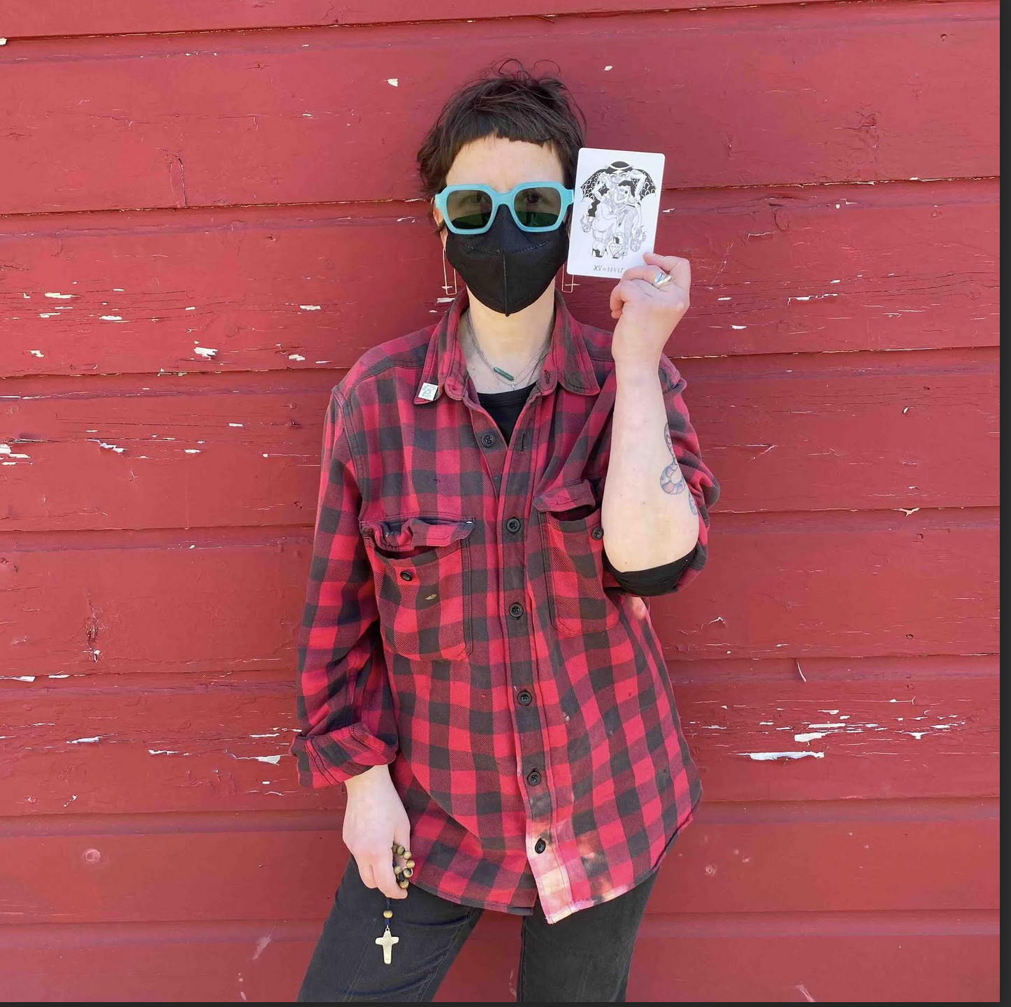 Person standing against a red wooden wall, wearing a black face mask, a large black and turquoise sunglasses, a red and black plaid shirt, and holding a tarot card in one hand.