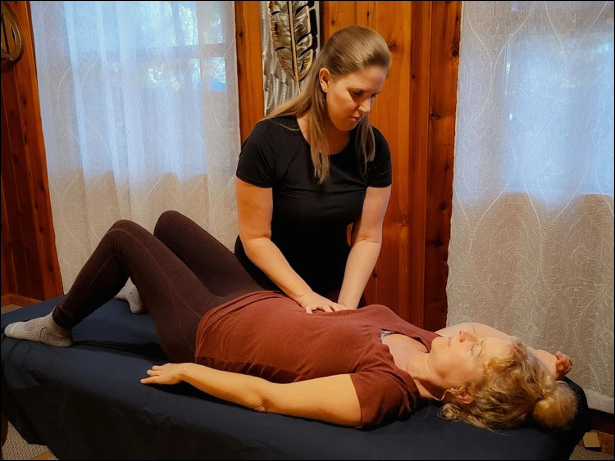 A woman giving a massage to another woman lying on a massage table in a cozy wood-paneled room.
