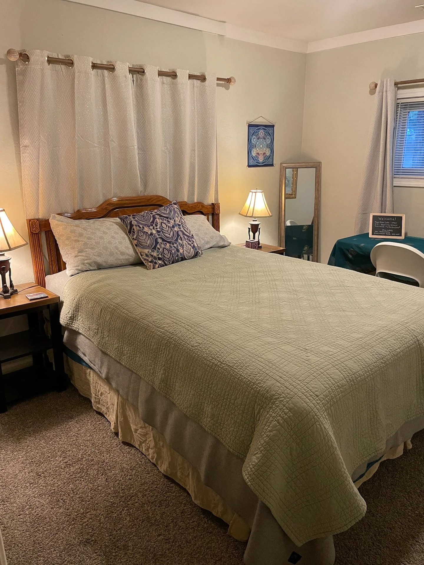 A cozy bedroom with a neatly made bed, beige quilt, and decorative pillows, flanked by two wooden nightstands each with a lamp, and a mirror leaning against the wall near a window with curtains.