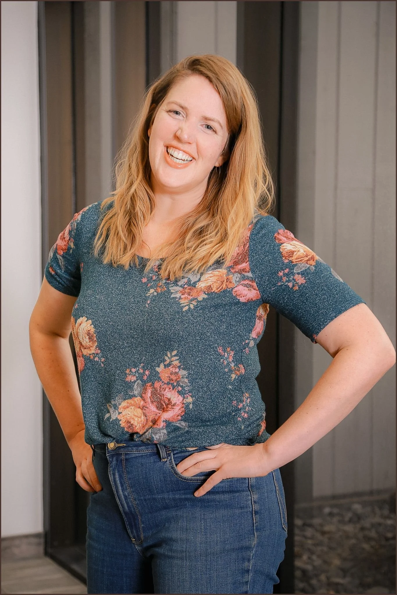 A woman with shoulder-length blonde hair, smiling and posing with her hands on her hips, wearing a blue floral shirt and blue jeans, standing indoors in front of a dark gray and black background.