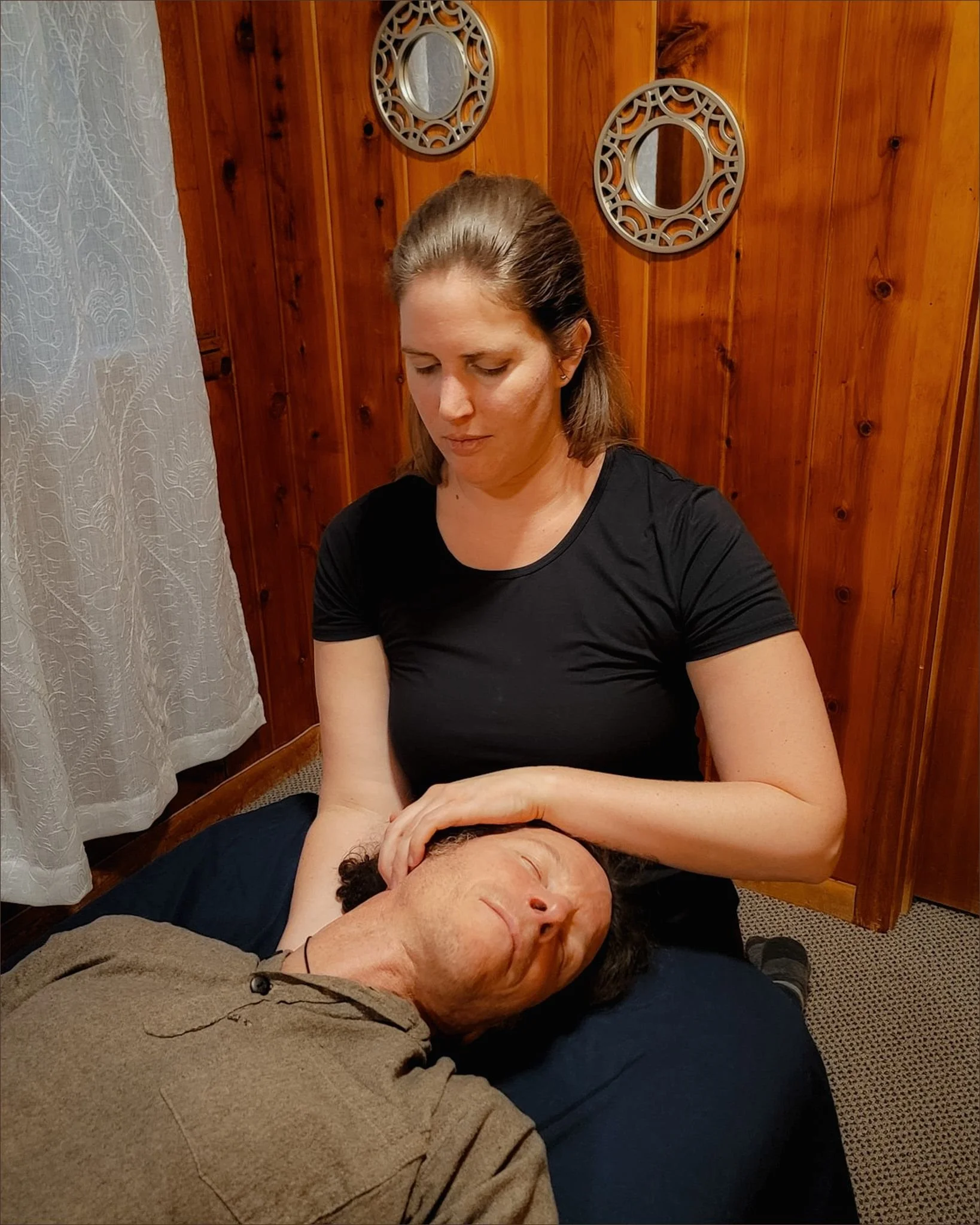 A woman providing a massage or therapy to a man lying on a treatment table in a room with wooden walls and decorative mirrors.