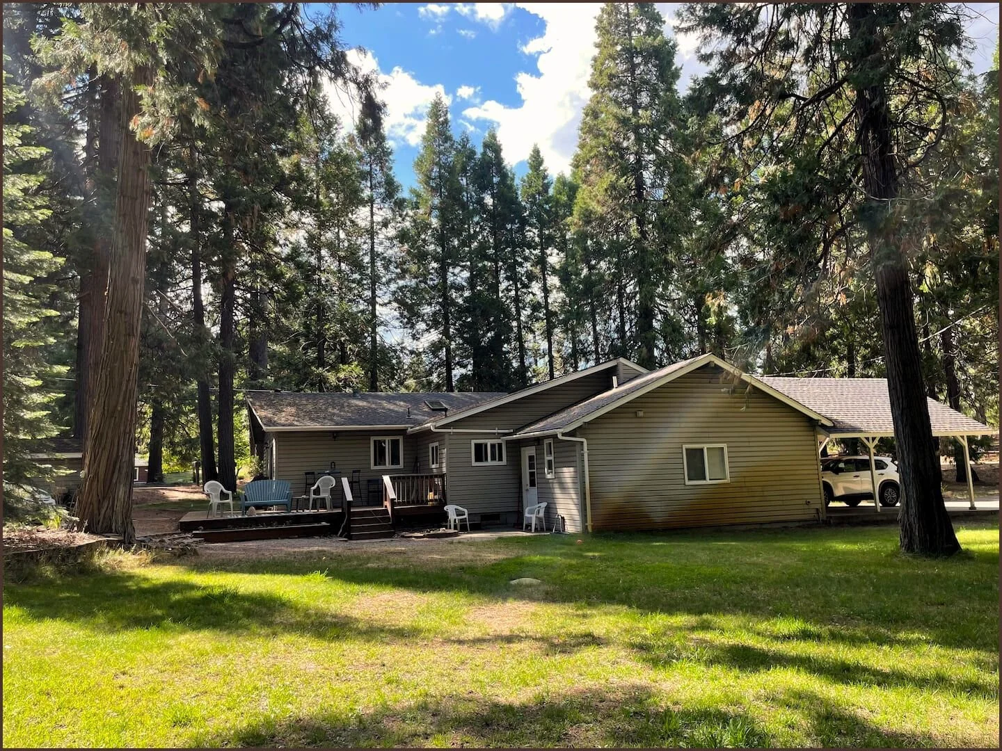A house in a wooded area with large trees surrounding it, a small deck with outdoor chairs, and a driveway with a parked white car. The house has beige siding and a sloped roof, with a grassy yard in the foreground.