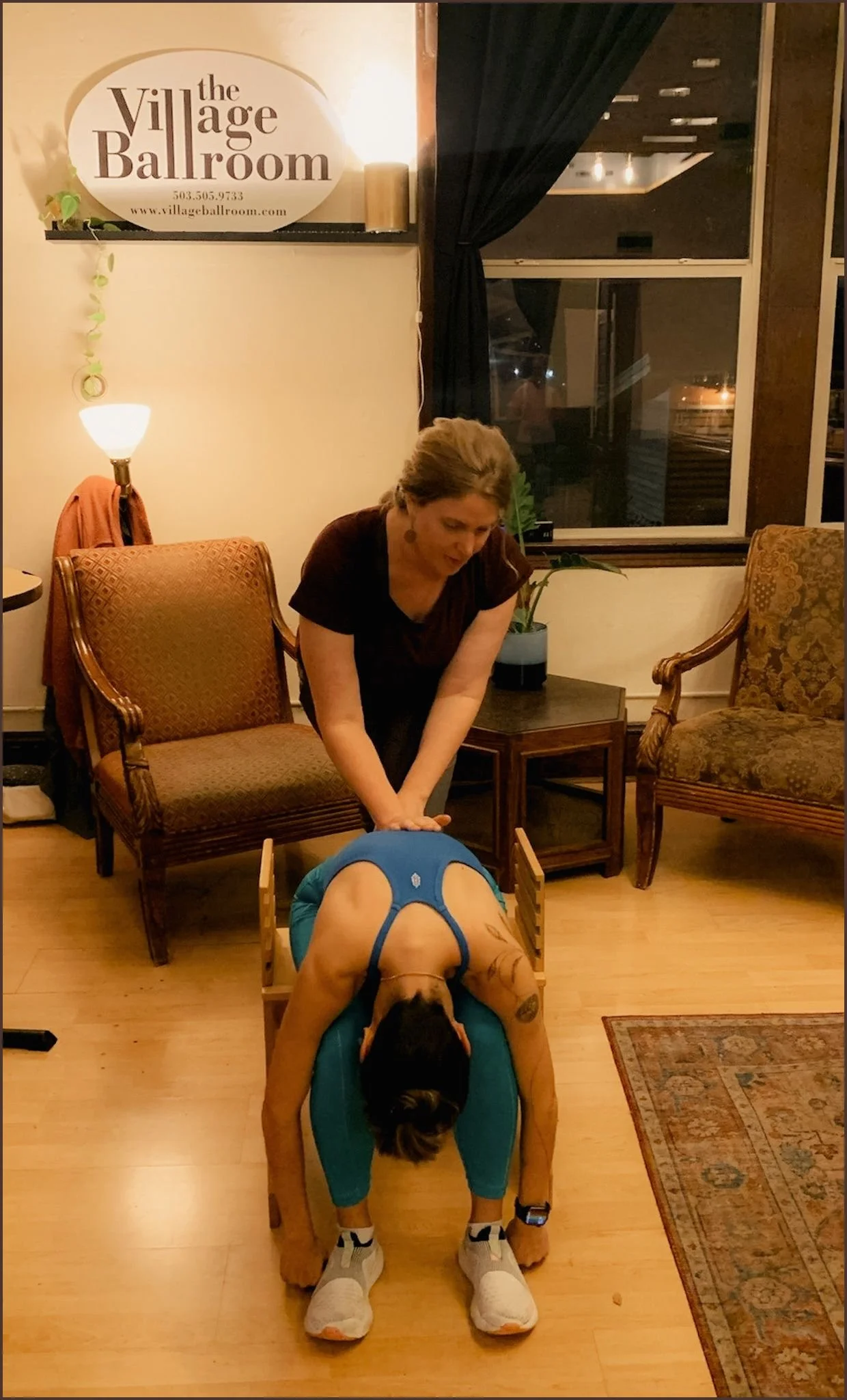 A woman giving a massage to a person lying face down on a massage table in a cozy room with vintage chairs and a window.