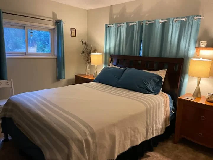 A bedroom with a large bed, dark wooden headboard, white and beige striped bedspread, and dark blue pillows. There are two wooden nightstands with lamps, one on each side of the bed. Blue curtains cover a window on one wall, and another window with a blue curtain is on an adjacent wall. A small decorative picture hangs on the wall near the window.