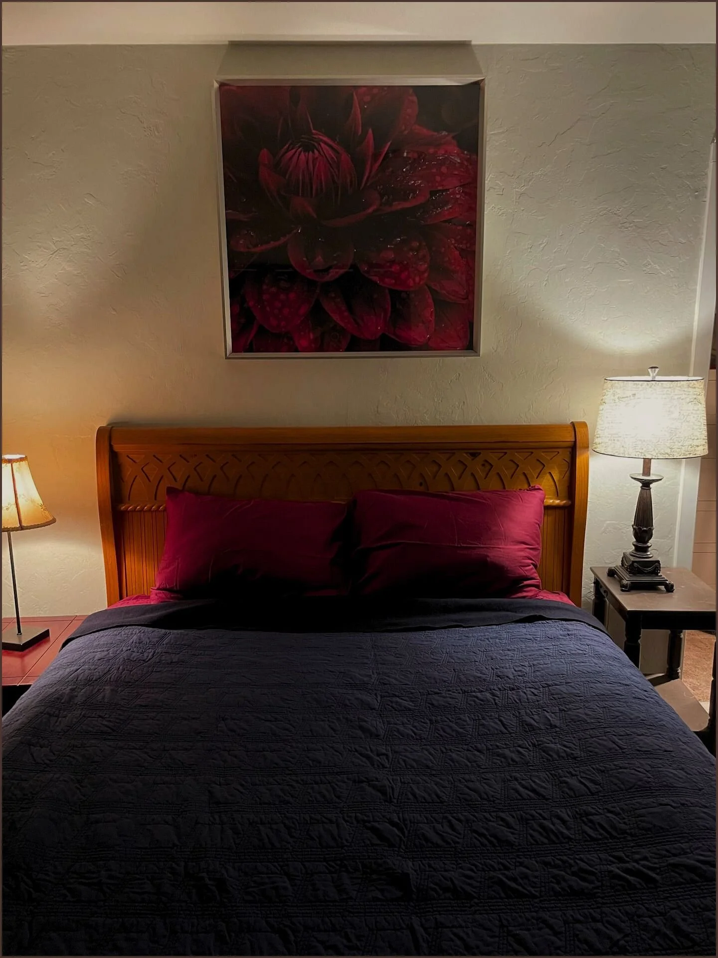 A bedroom with a wooden bed frame, a dark quilt, and two red pillows. There are two bedside lamps with cream-colored lampshades on black side tables. A framed flower photograph hangs above the bed.