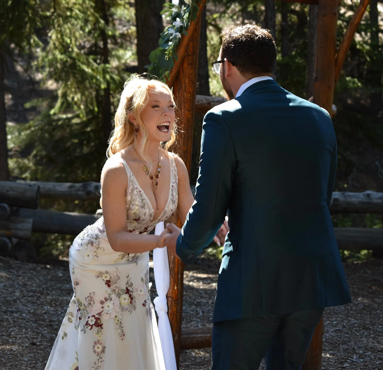 A bride at a wedding outdoors, holding hands and laughing. They are under a wooden beam at Skyliners Lodge in Central Oregon