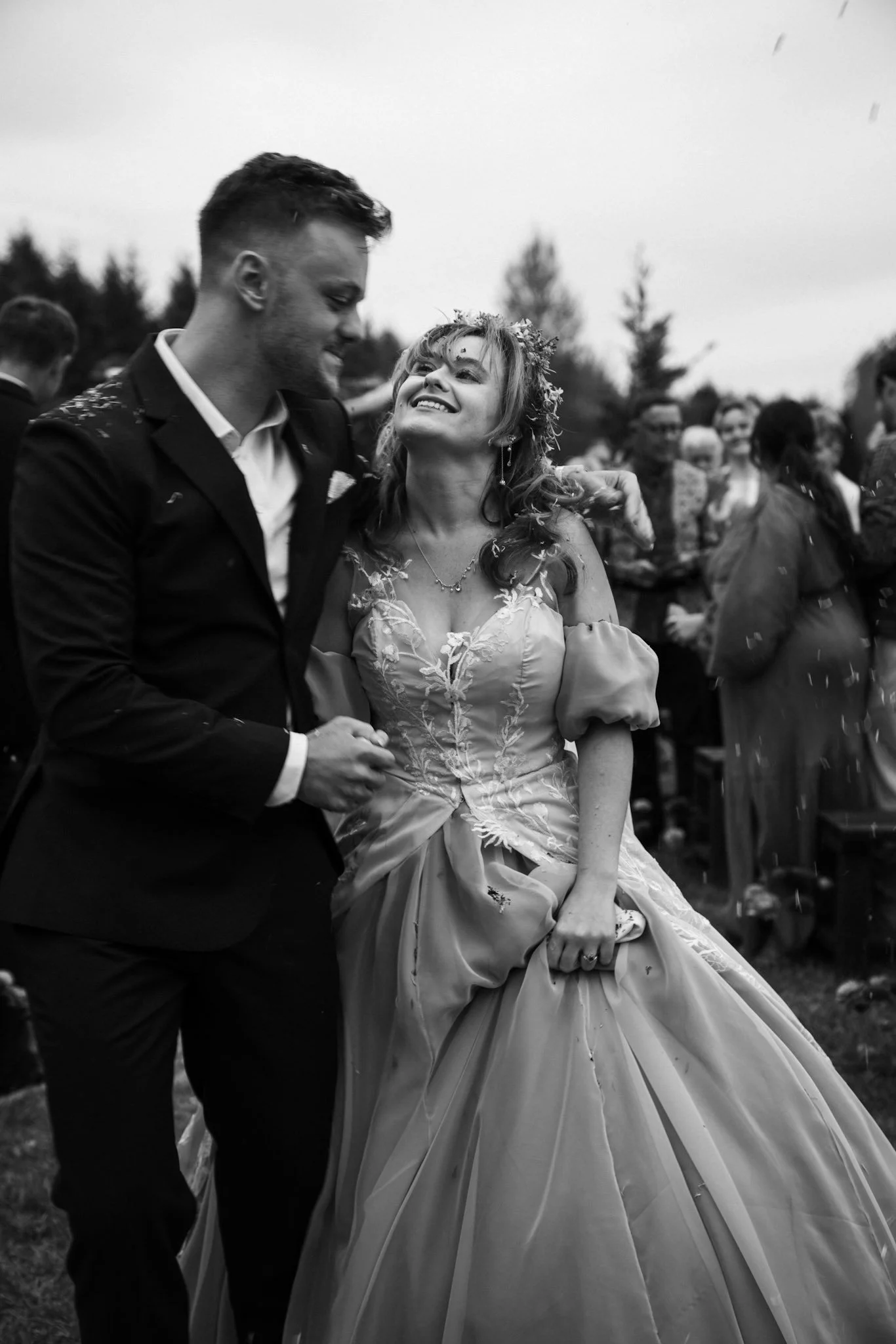 Black and white photo of a couple dancing at a wedding, the woman wearing a wedding dress and crown, smiling, with guests in the background.