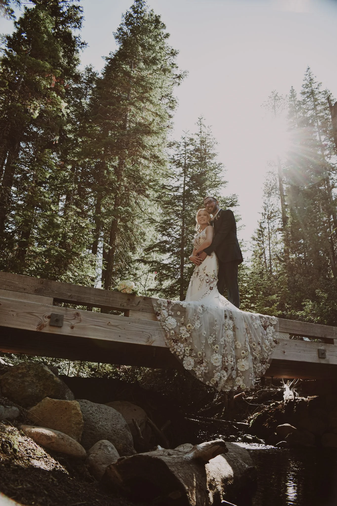 A bride and groom in wedding attire standing on a wooden bridge in a forest, with sunlight shining through trees behind them.