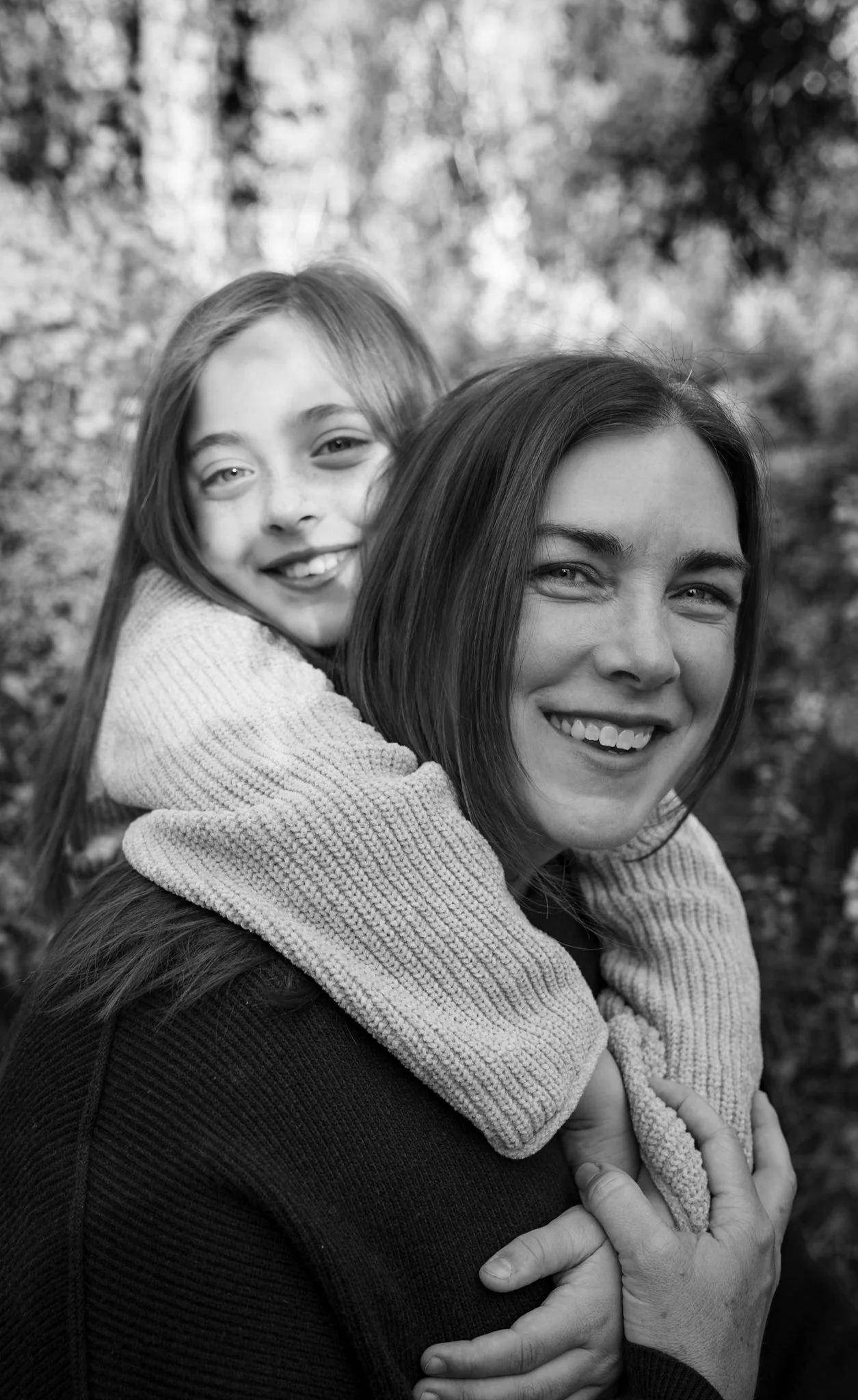 A woman with shoulder-length hair smiling, with a young girl wearing a sweater hugging her from behind, outdoors with blurred trees in the background.