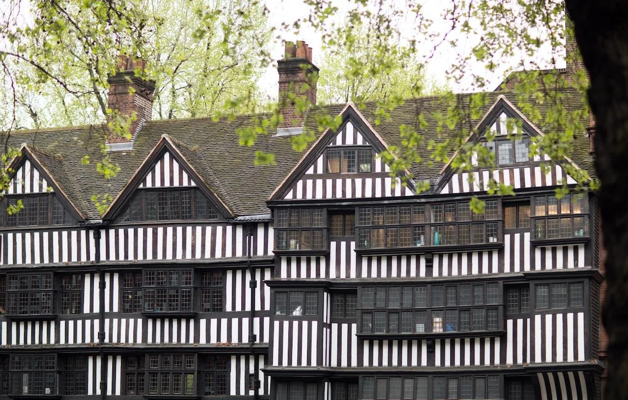A large multi-story building with a black and white striped Tudor-style façade, surrounded by tall trees with green leaves.