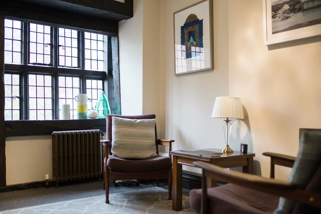 Cozy corner of a therapy room with two armchairs, a side table with a lamp, and a large window with decorative vases on the sill. Artwork hangs on the wall.