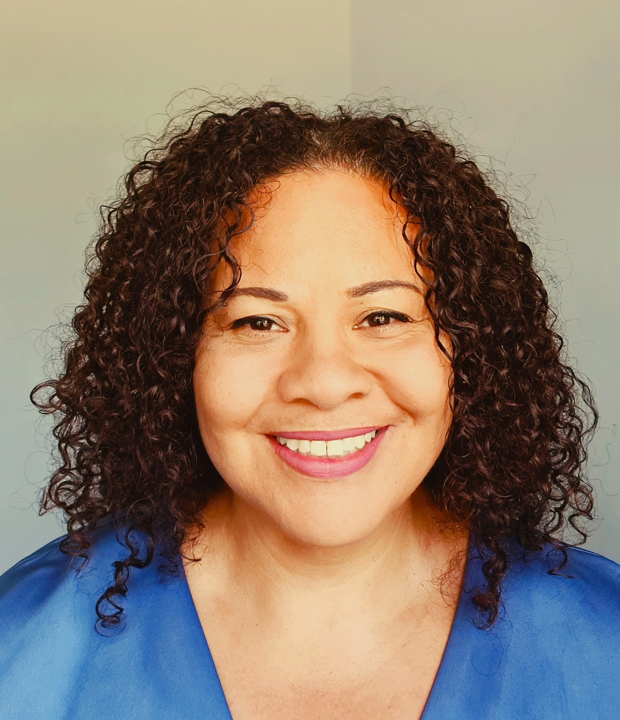 Close-up photo of a woman with curly dark hair, smiling, wearing a blue top, against a plain gray background.
