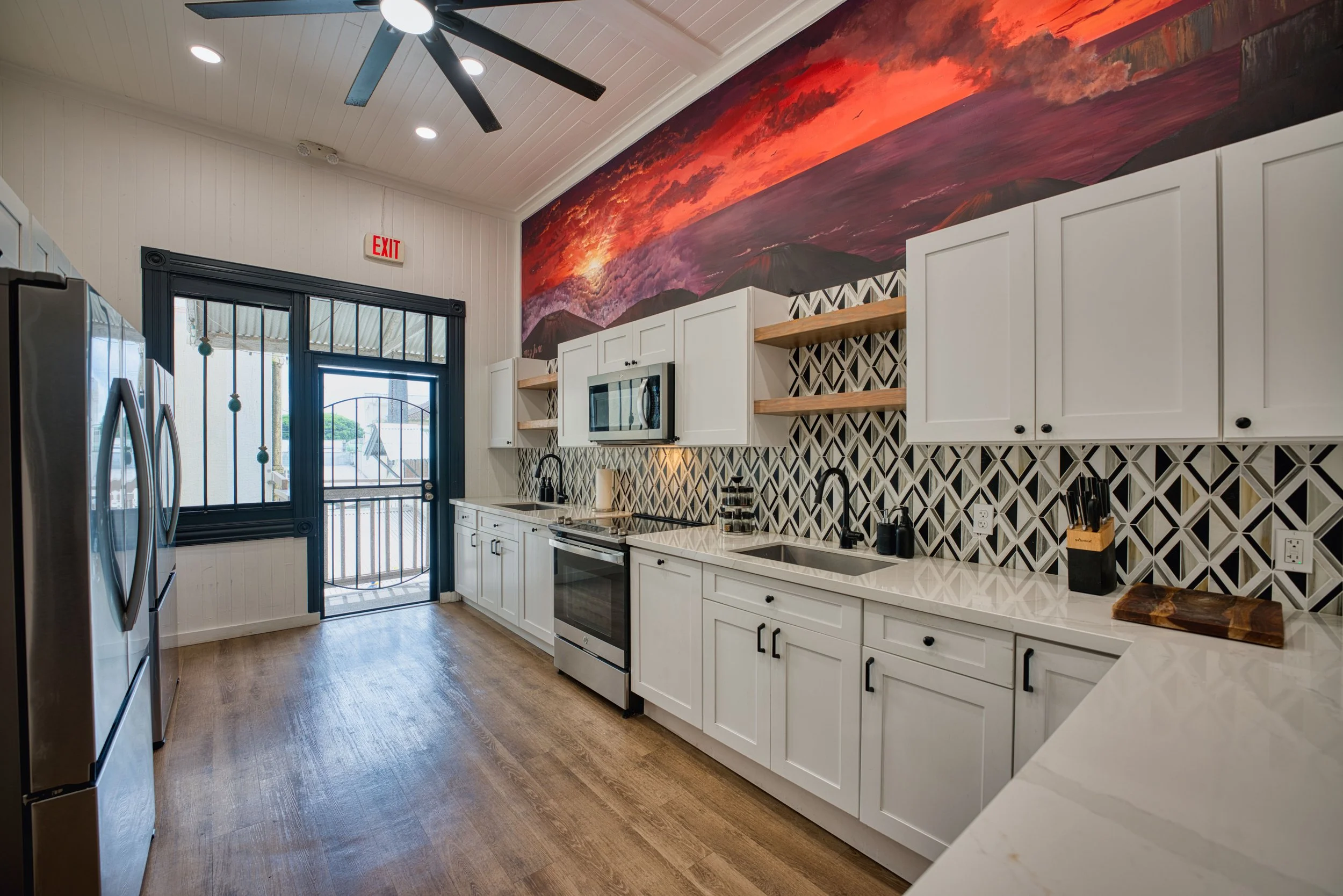 Modern kitchen with white cabinets, black hardware, geometric tile backsplash, wooden open shelves, and a mural of a sunset or sunrise above the cabinets.