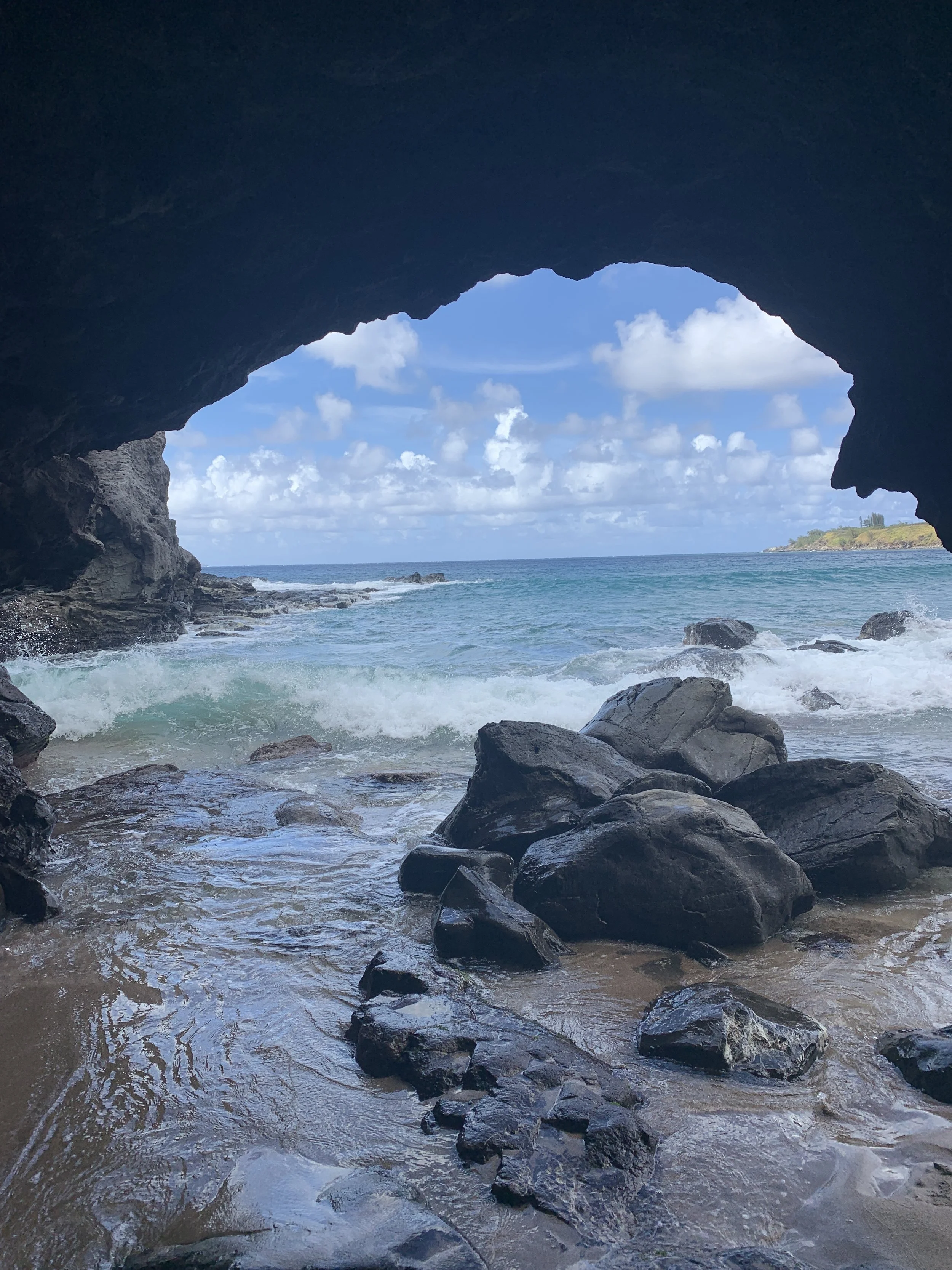 View from inside a rocky cave looking out at ocean waves and a partly cloudy sky, with a distant coastline.