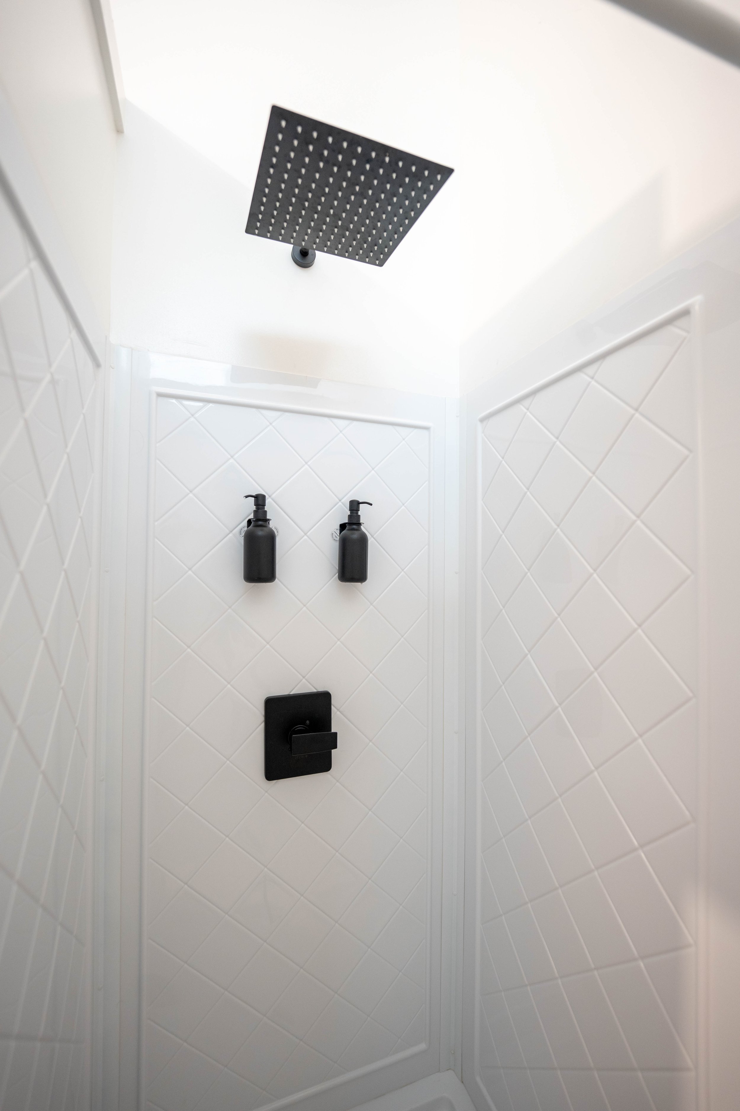 Shower interior with black showerhead, two black soap dispensers, and black shower control on a white tiled wall.