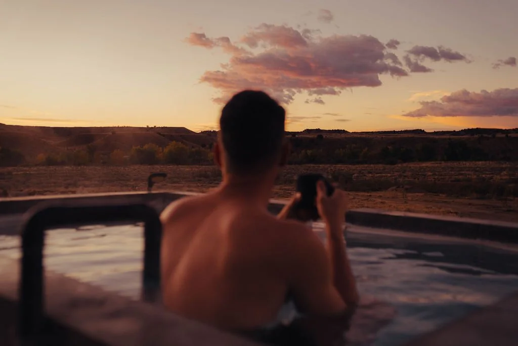 A person relaxing in a hot tub outdoors during sunset, taking a photo of the colorful sky with clouds and a distant horizon.