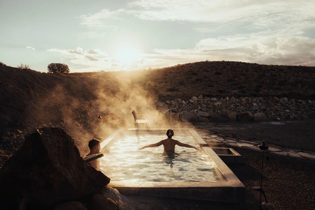 People soaking in an outdoor hot spring during sunset, surrounded by rocky terrain and open sky.
