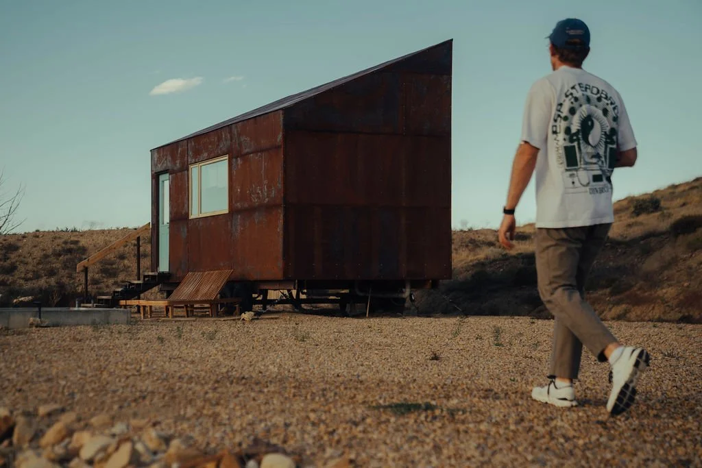 Rustic tiny house on wheels in a desert landscape with a small swimming pool in the foreground.