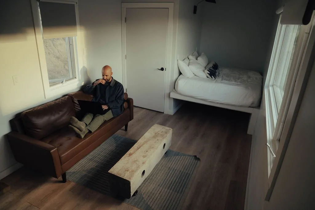 A man sitting on a brown leather couch working on a laptop, with a bed in the corner of the room, and windows allowing natural light to enter.