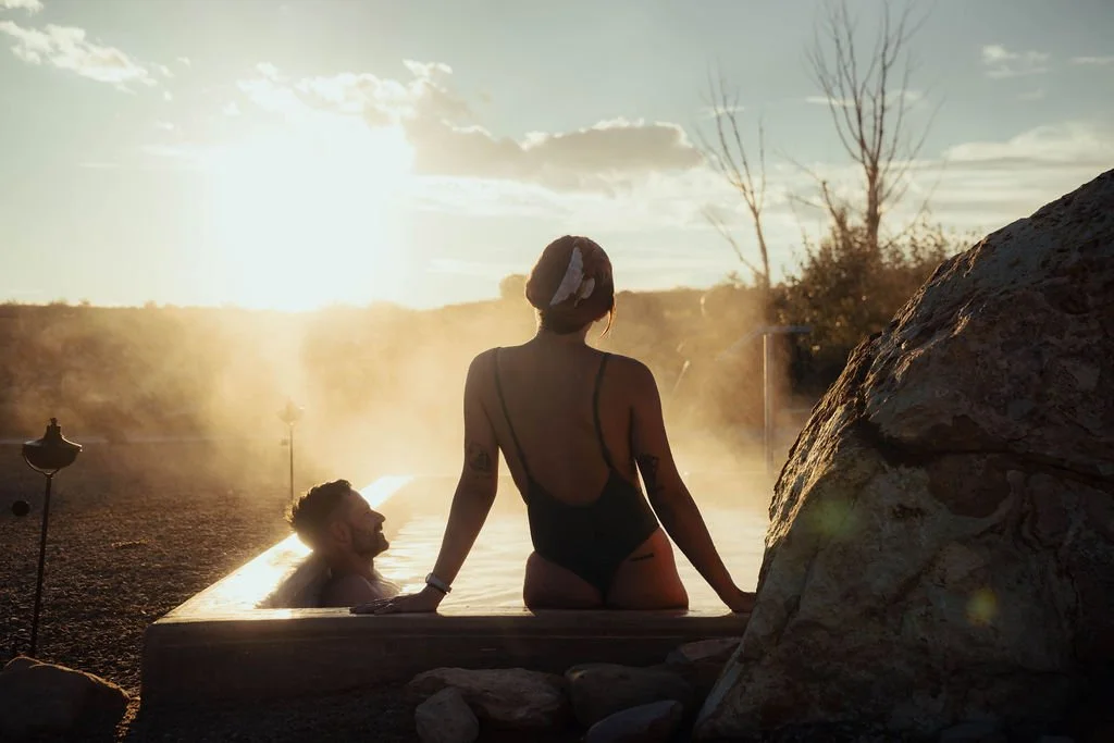 A man and a woman in an outdoor hot spring at sunset, surrounded by rocks and landscape with barren trees and a cloudy sky.