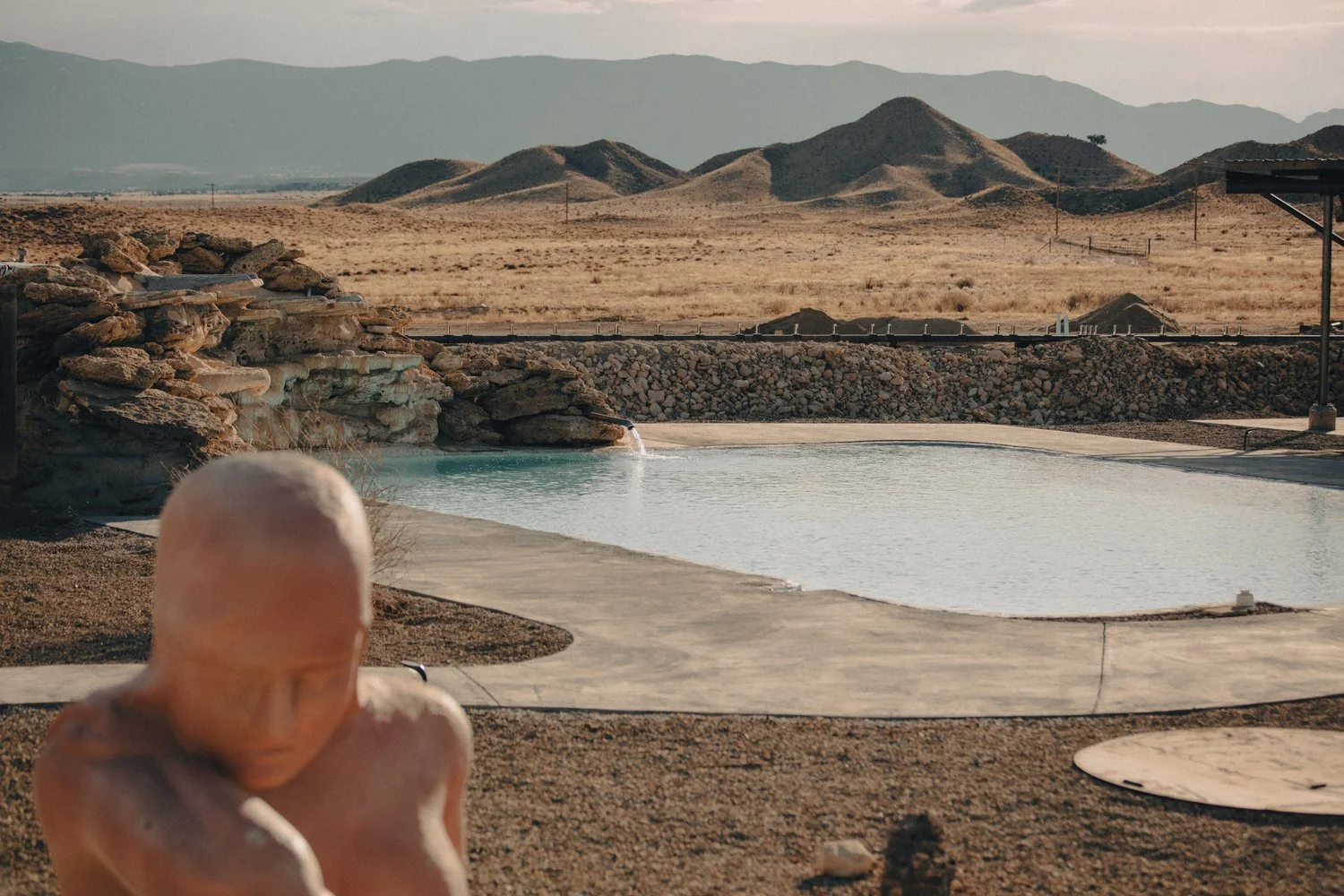 A hot spring in a desert landscape with barren hills and mountains in the background, and a small rocky waterfall flowing into the pool in the foreground.