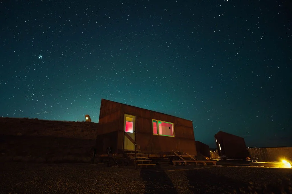 A small modern cabin with colorful lit windows, set against a starry night sky on a hillside.