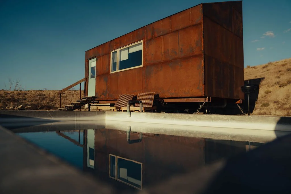 A small, rusty metal tiny house on wheels with a white door and two windows, outdoor wooden stairs, and a seating area with two chairs in a desert landscape under a blue sky.