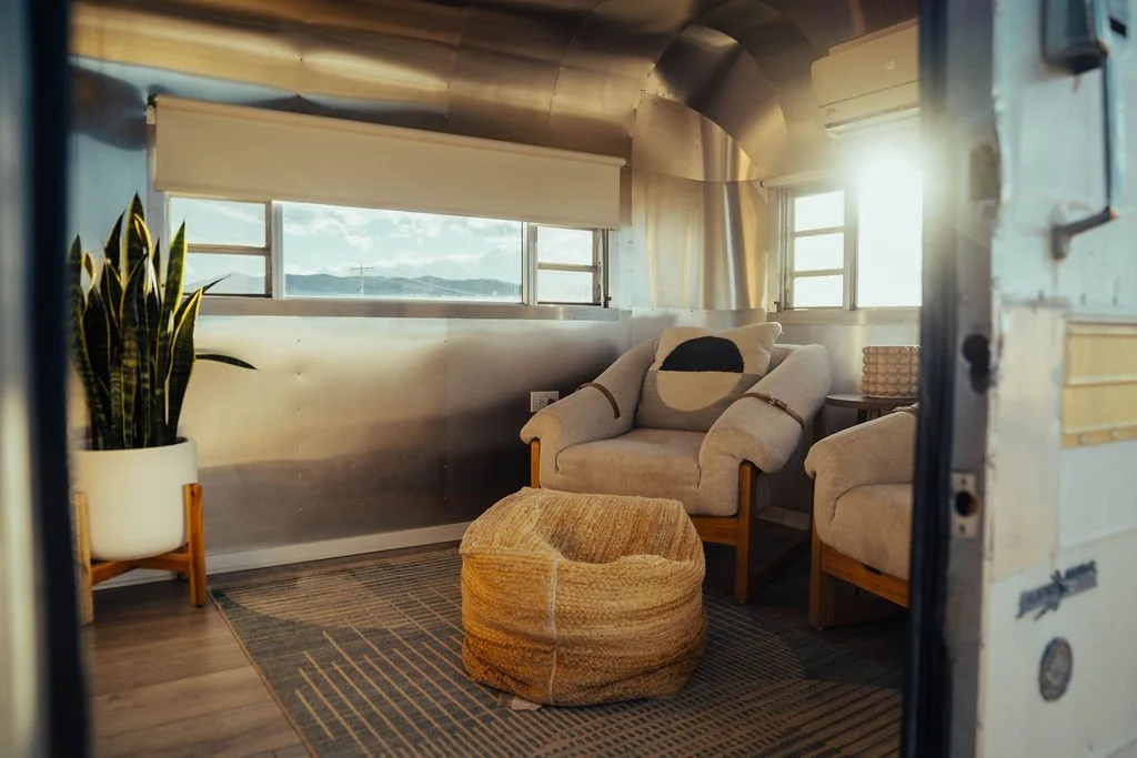 Interior of a cozy living space inside a renovated vintage trailer with a large window, a comfortable armchair with a pillow, a potted plant, a woven ottoman, and decorative elements.