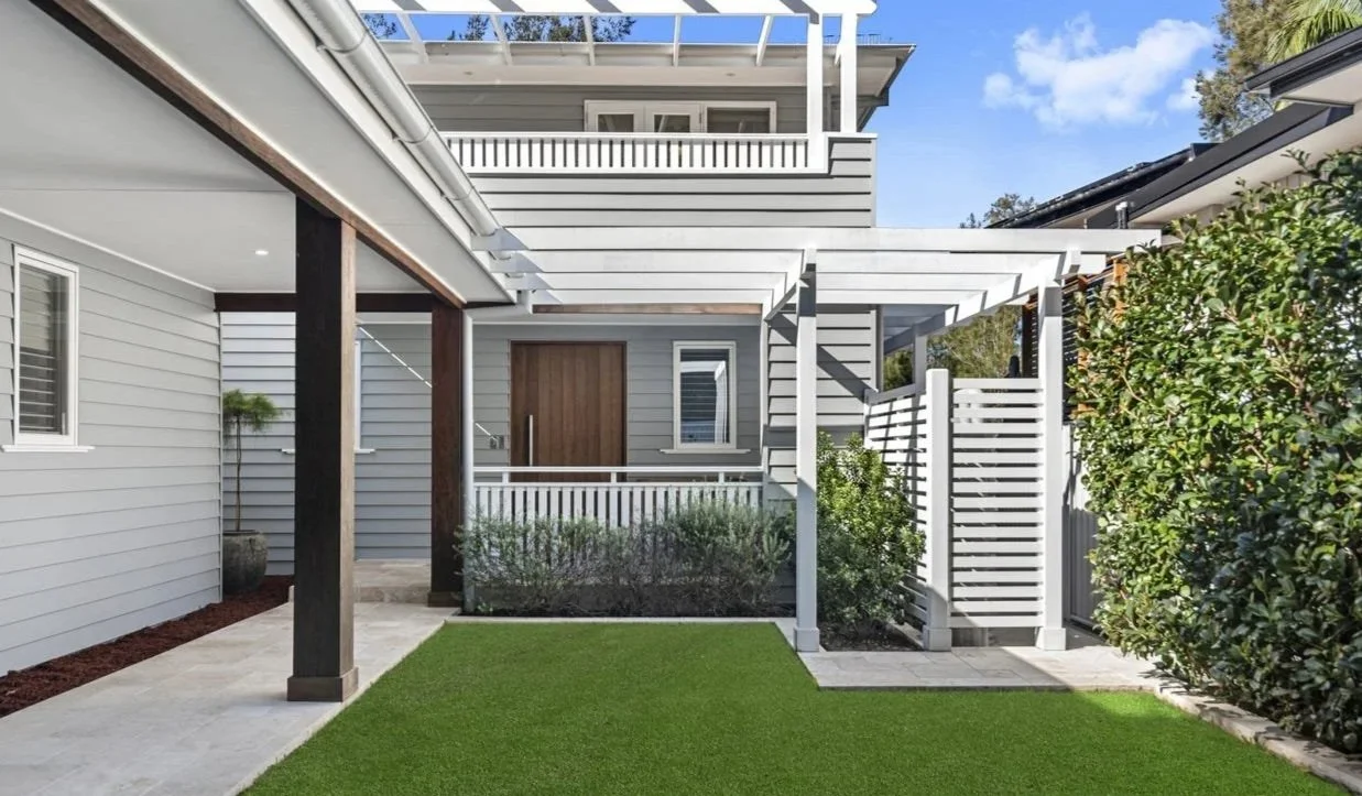 Modern backyard with gray house, white fencing, pergola, artificial grass, potted plant, and surrounding greenery.